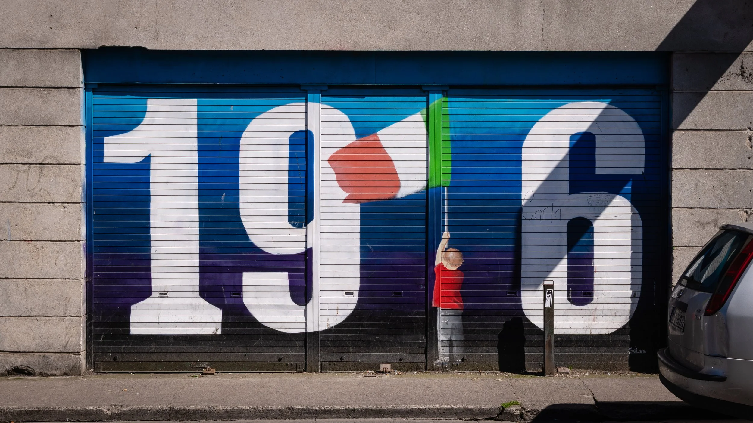 Garage door painted with large numbers '10' and '6' in white, with colorful abstract shapes, a person in a red shirt standing in front, and parts of a car visible on the right.
