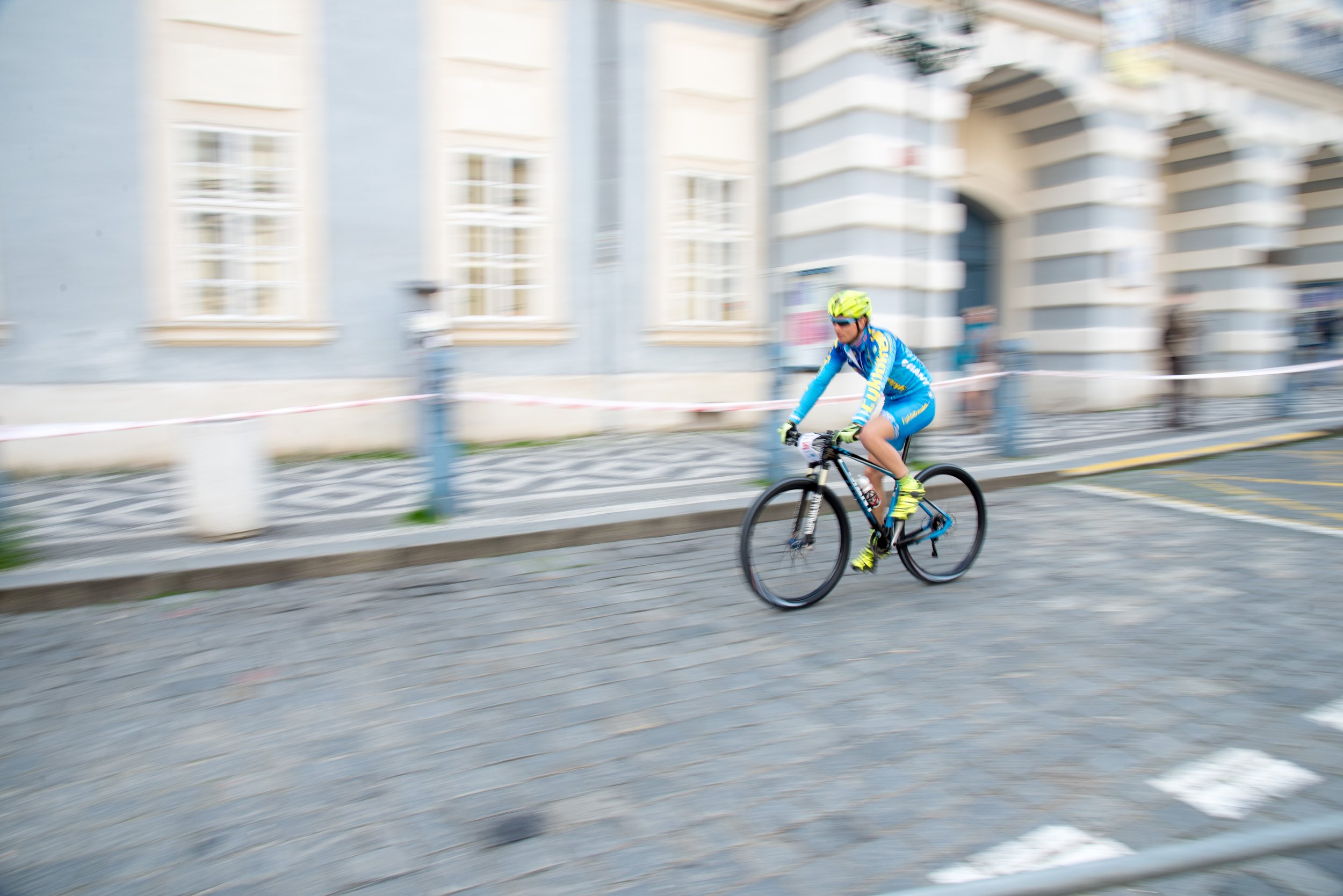 Cyclist in bright blue and yellow gear riding a mountain bike on cobblestone street, with blurred city buildings and spectators in background.