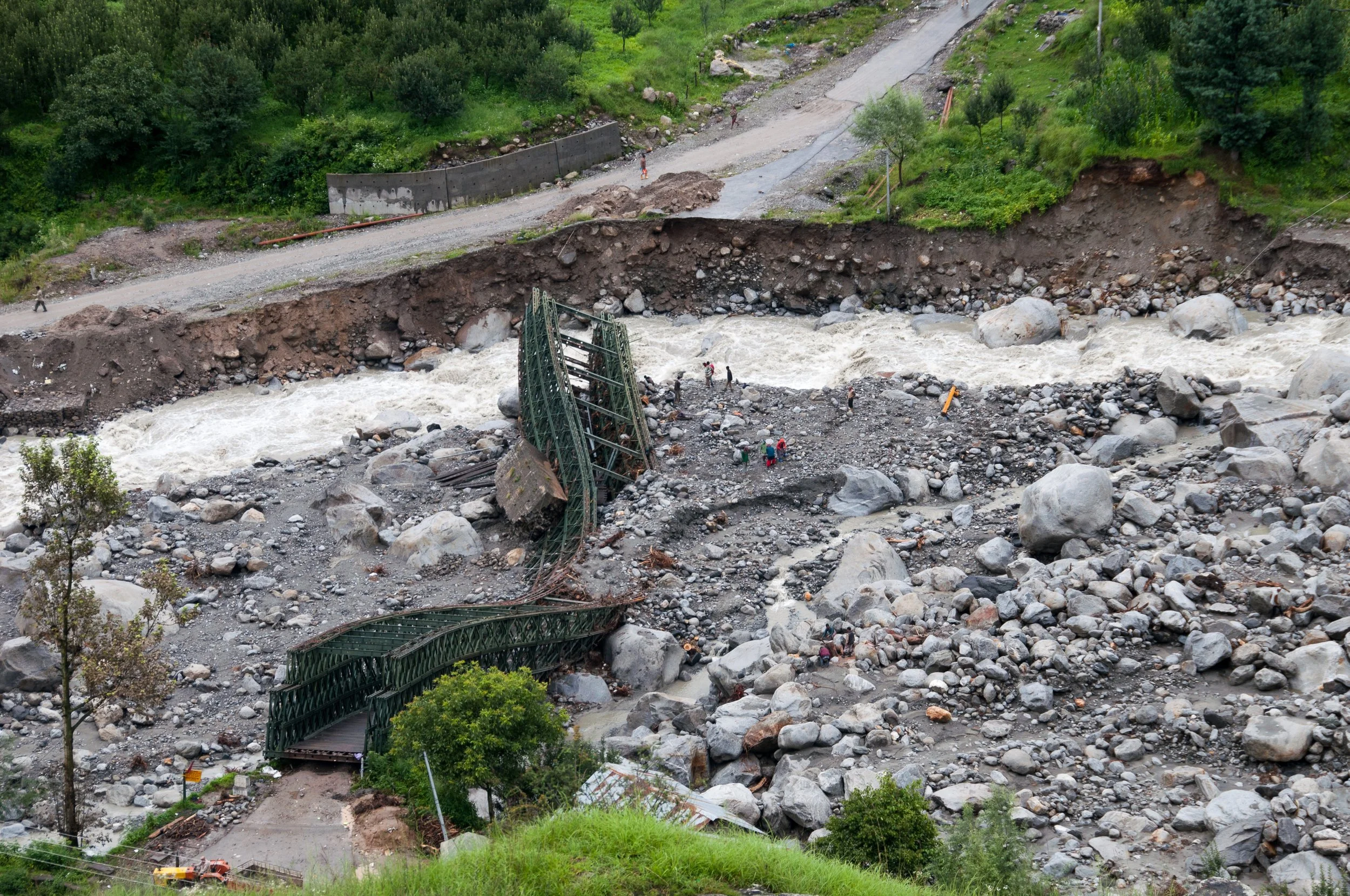 Damaged green bridge over rocky riverbed caused by landslide with scattered rocks and debris, with some people visible exploring area