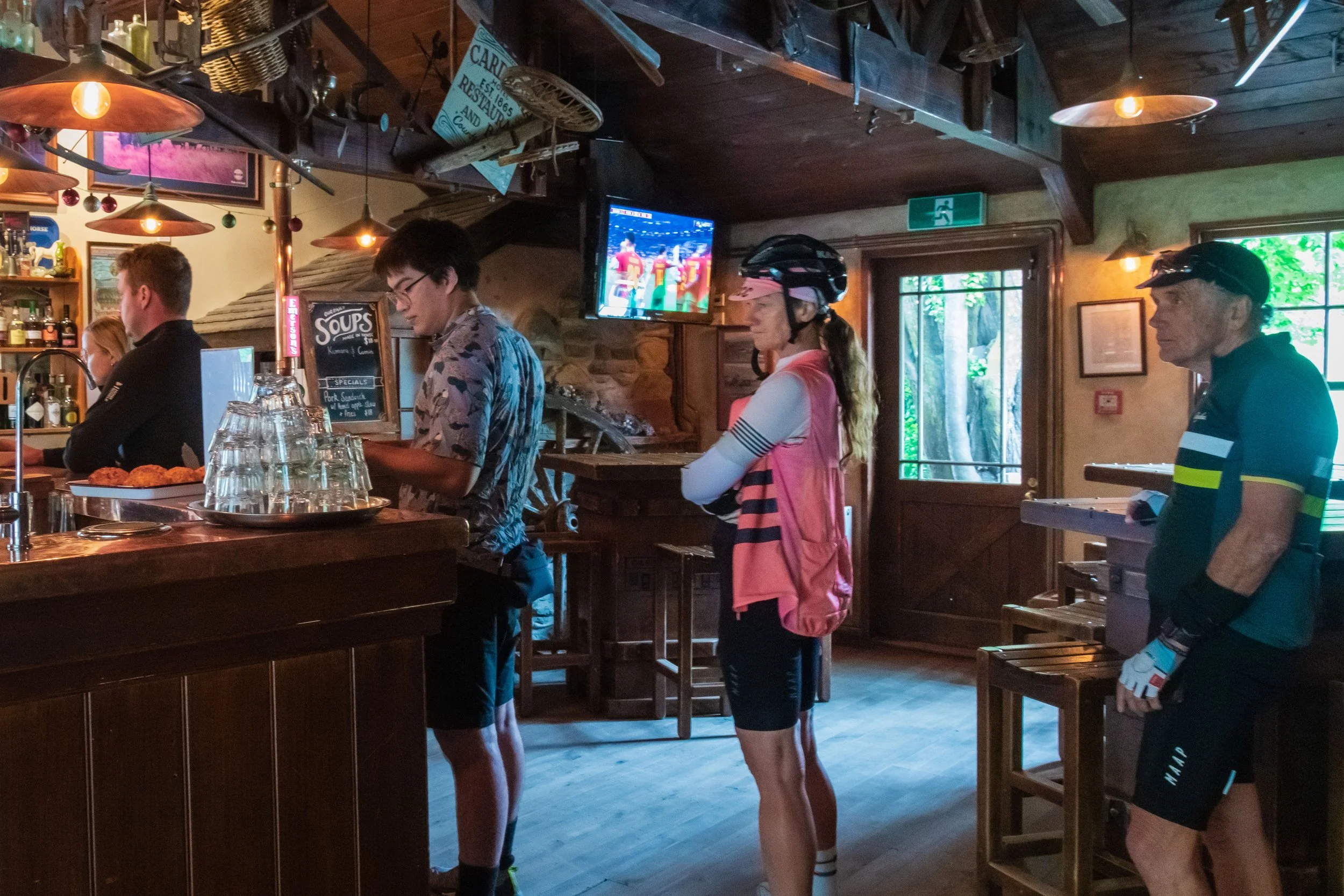 People waiting in line inside a rustic bar or restaurant, with a bartender behind the counter and a television mounted on the wall.