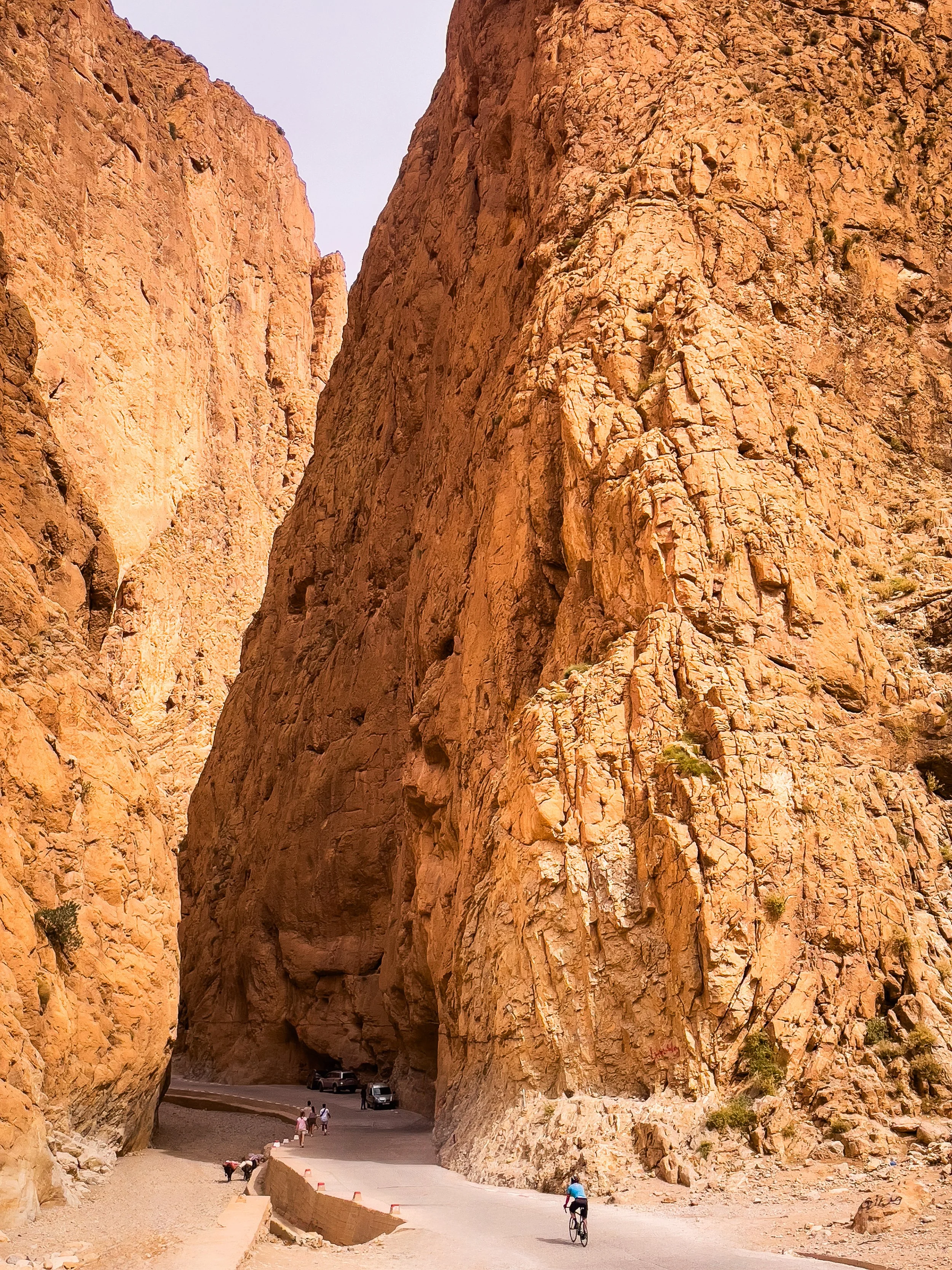 A narrow mountain pass with tall, reddish-orange rocky cliffs on both sides and a paved road running through it. There are a few people, including a cyclist and pedestrians, along the road with some vehicles parked at the far end.