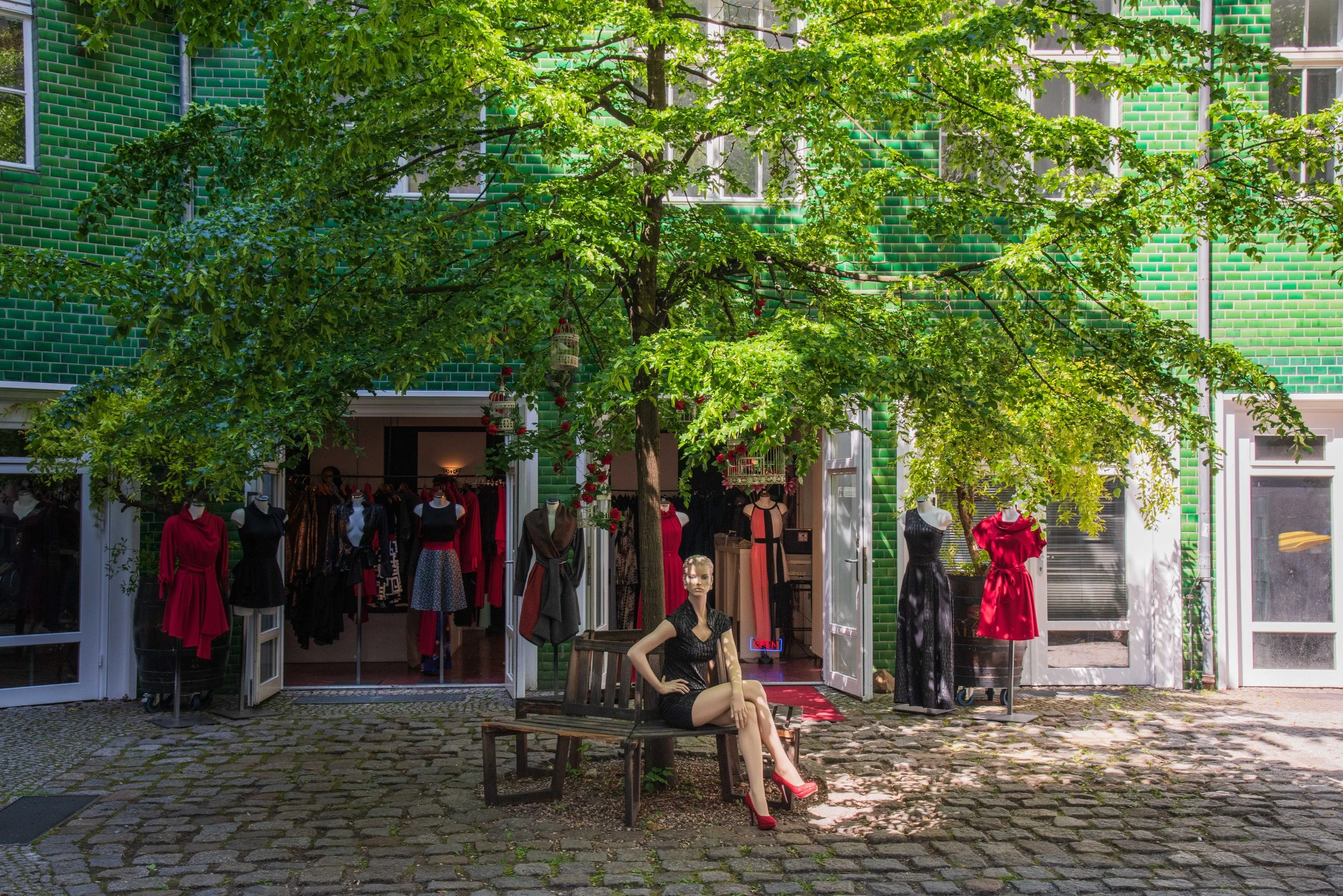 A clothing boutique with an open front displaying various dresses, with a mannequin sitting on a bench outside wearing a black dress and red high heels, under a leafy green tree on a cobblestone courtyard.