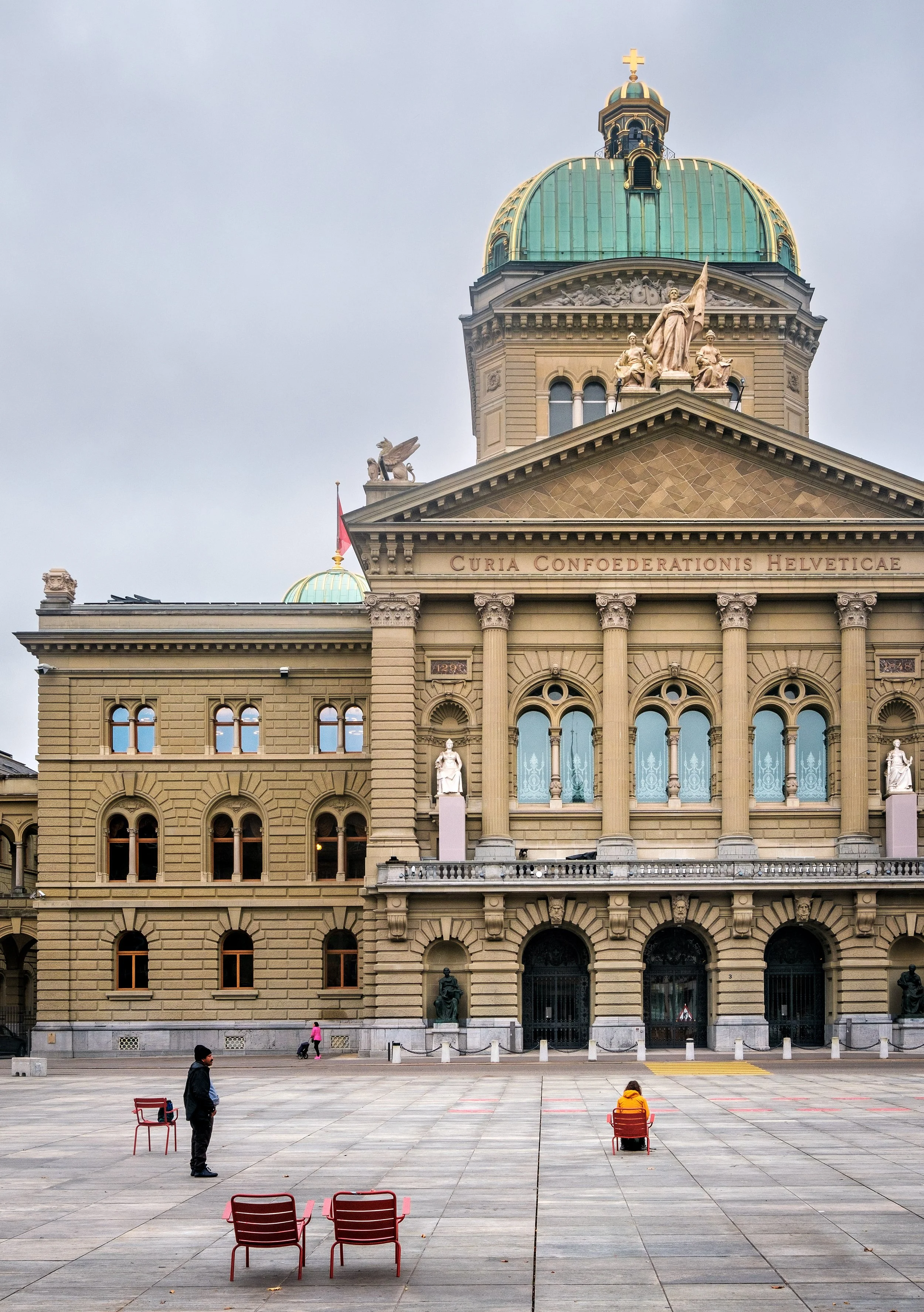 View of a historic building with a green dome and beige stone exterior, featuring statues and architectural details, with several people sitting and standing in the open plaza in front.