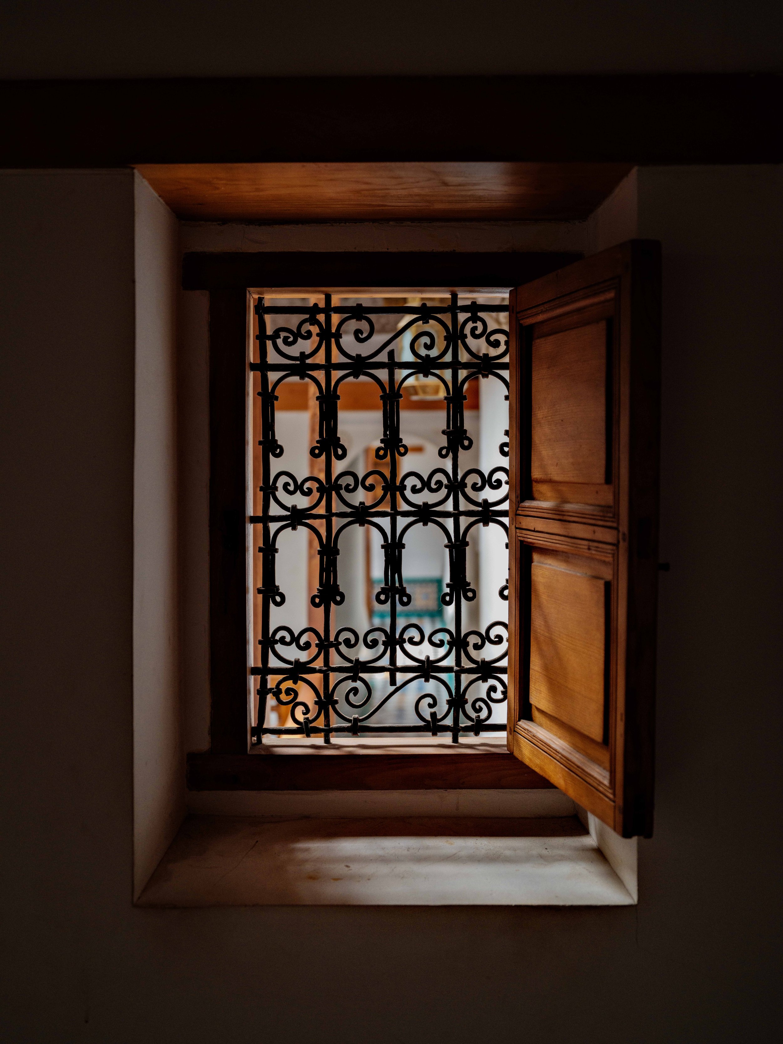 View through a small open window with decorative wrought iron bars and a wooden shutter on one side, showing an interior courtyard or building with wooden beams and white walls.