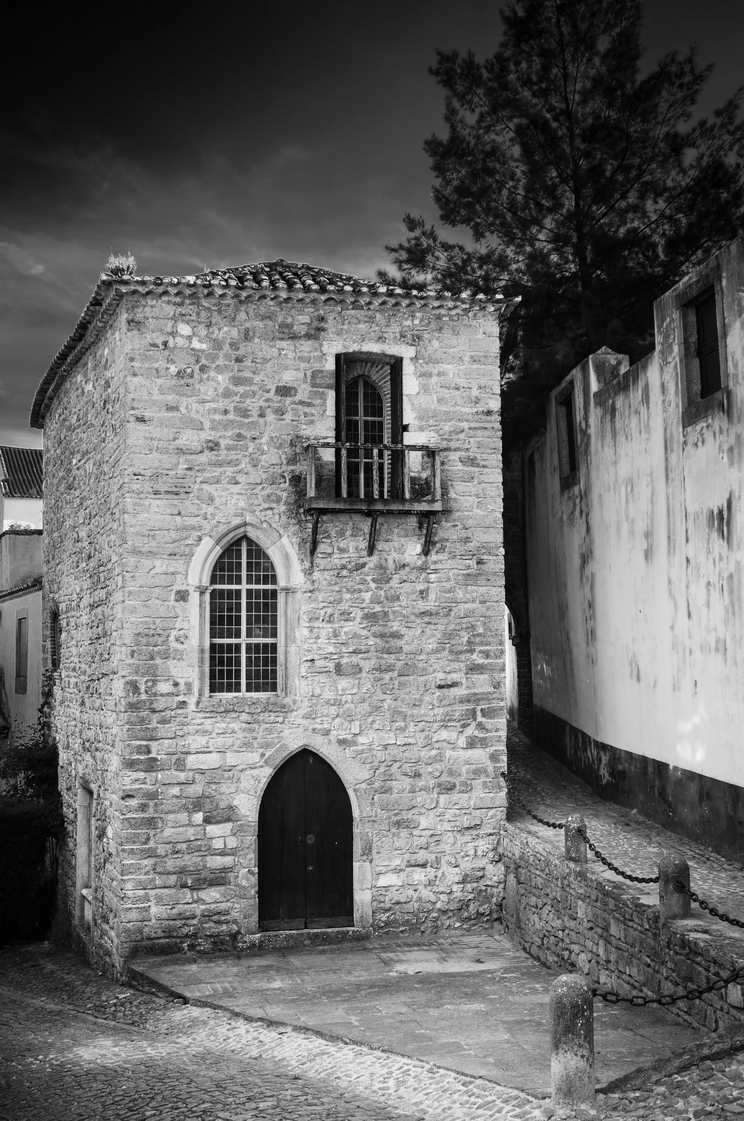 Black and white photo of a small, old stone building with arched windows, a wooden door, and a small balcony, surrounded by a cobblestone street and a chain-linked barrier.