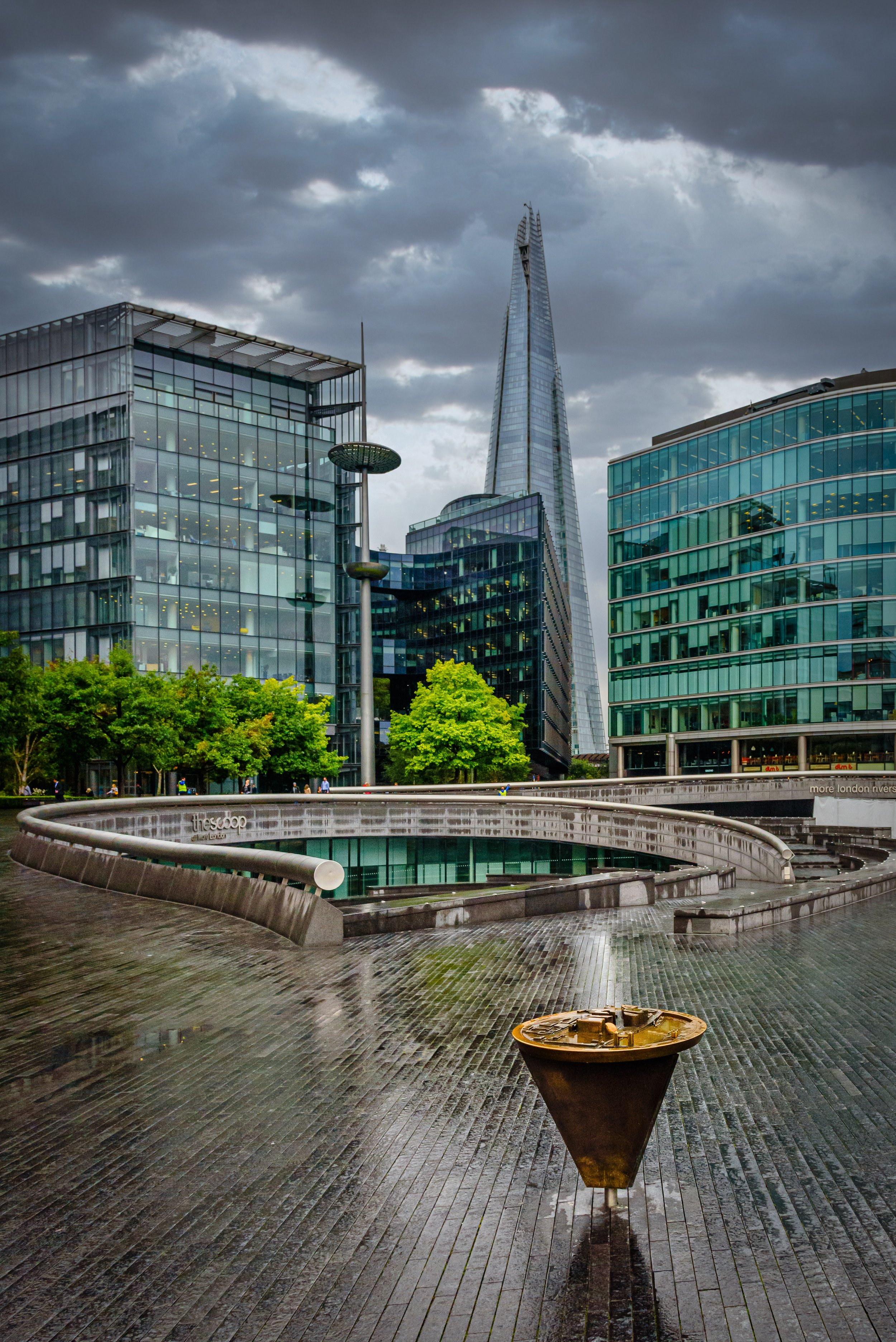 Cityscape with modern glass buildings, the Shard in London, and a water feature in the foreground on a rainy day with dark clouds.