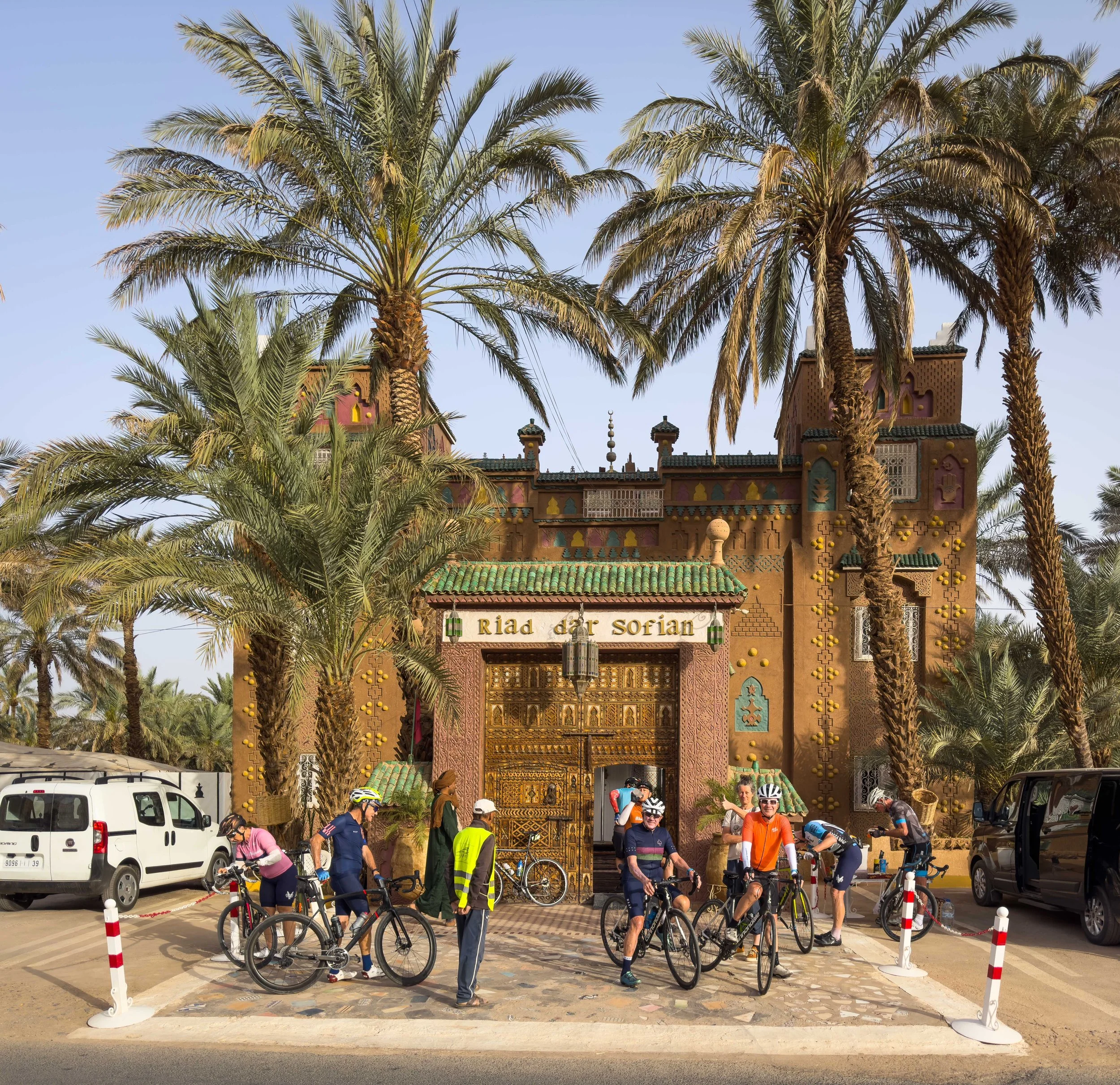 Group of cyclists in front of elaborate Moroccan-style gate with palm trees and vehicles nearby, at the entrance of Riad Dar Sofian.