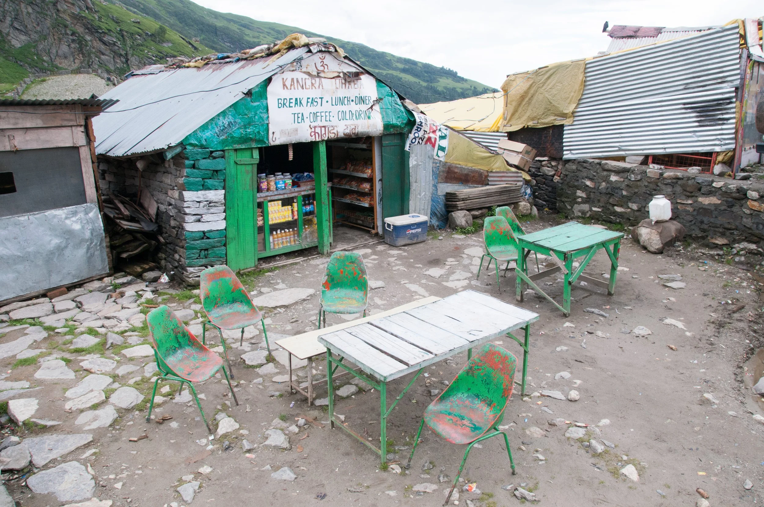 Small roadside eatery with a corrugated metal roof, painted green doors, and a sign offering breakfast, lunch, dinner, tea, coffee, and cold drinks. There are four green metal chairs and two tables outside on a dirt and stone ground. In the backgroun