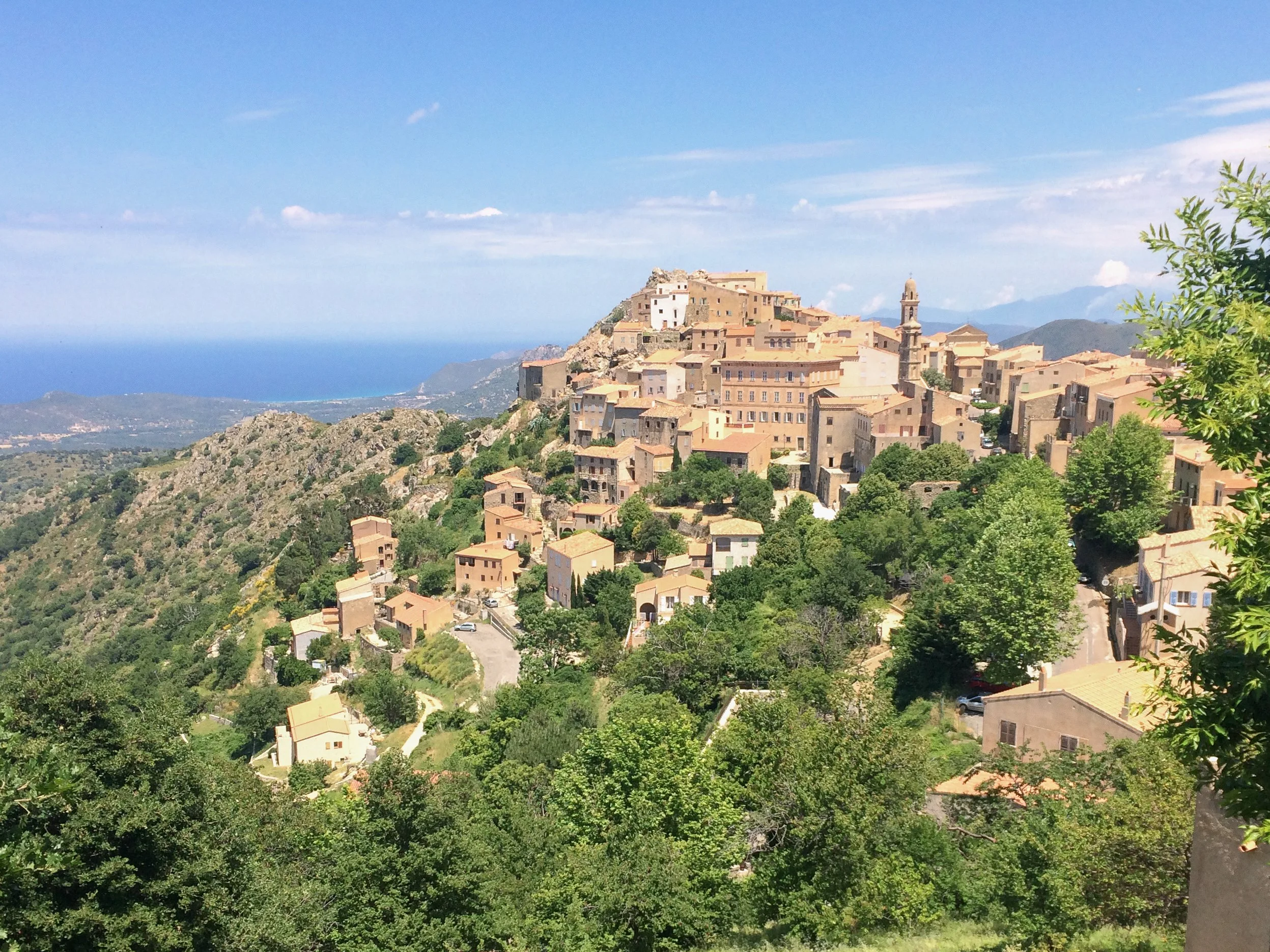 View of a hillside town with beige buildings and a church tower, surrounded by green trees, overlooking the sea in the distance under a blue sky with some clouds.