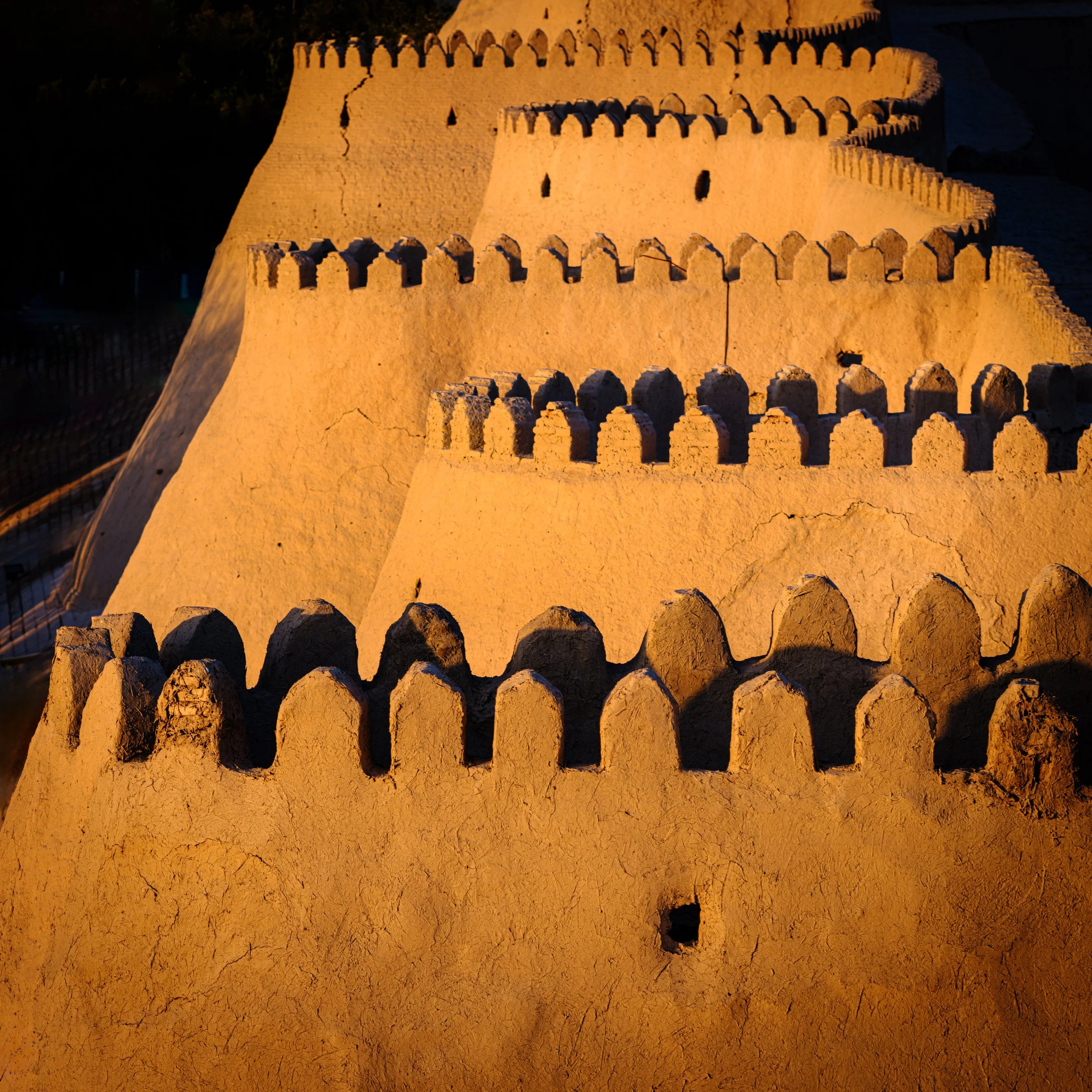 Close-up of an adobe fortress wall with crenellations, lit by warm light against a dark background.