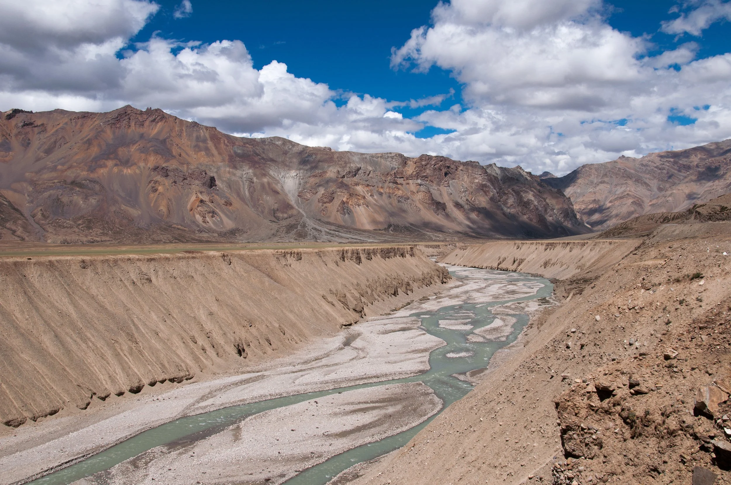A dry, rugged canyon with steep banks and a winding river running through it, set against distant mountains and a partly cloudy sky.