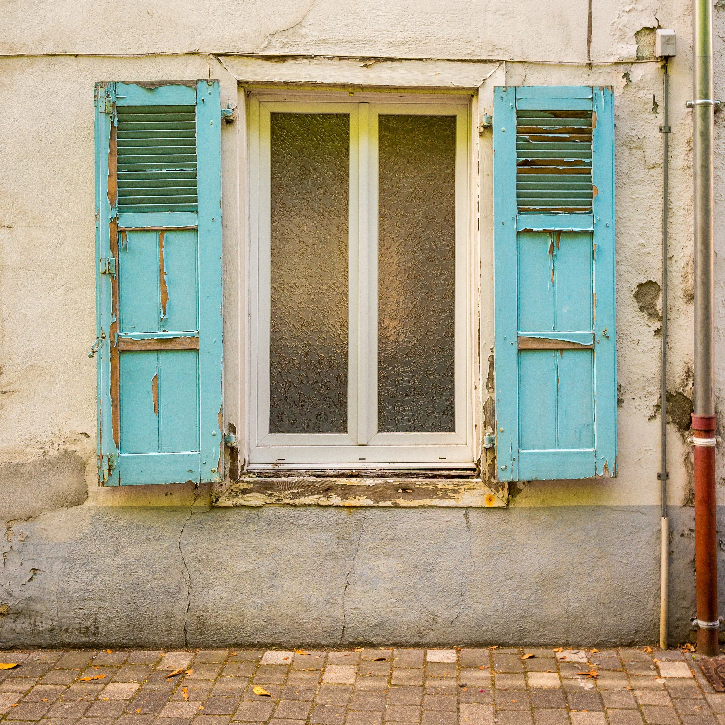 A window with blue peeling shutters on a cracked and weathered beige wall, with a cobblestone sidewalk in front.