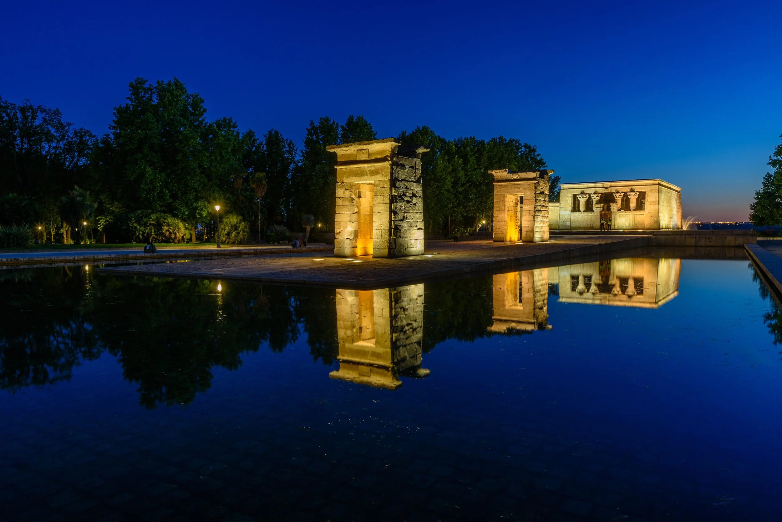 Night view of ancient Egyptian temples illuminated by lights, reflected in a still water pool. Tall trees and a darkening sky in the background.