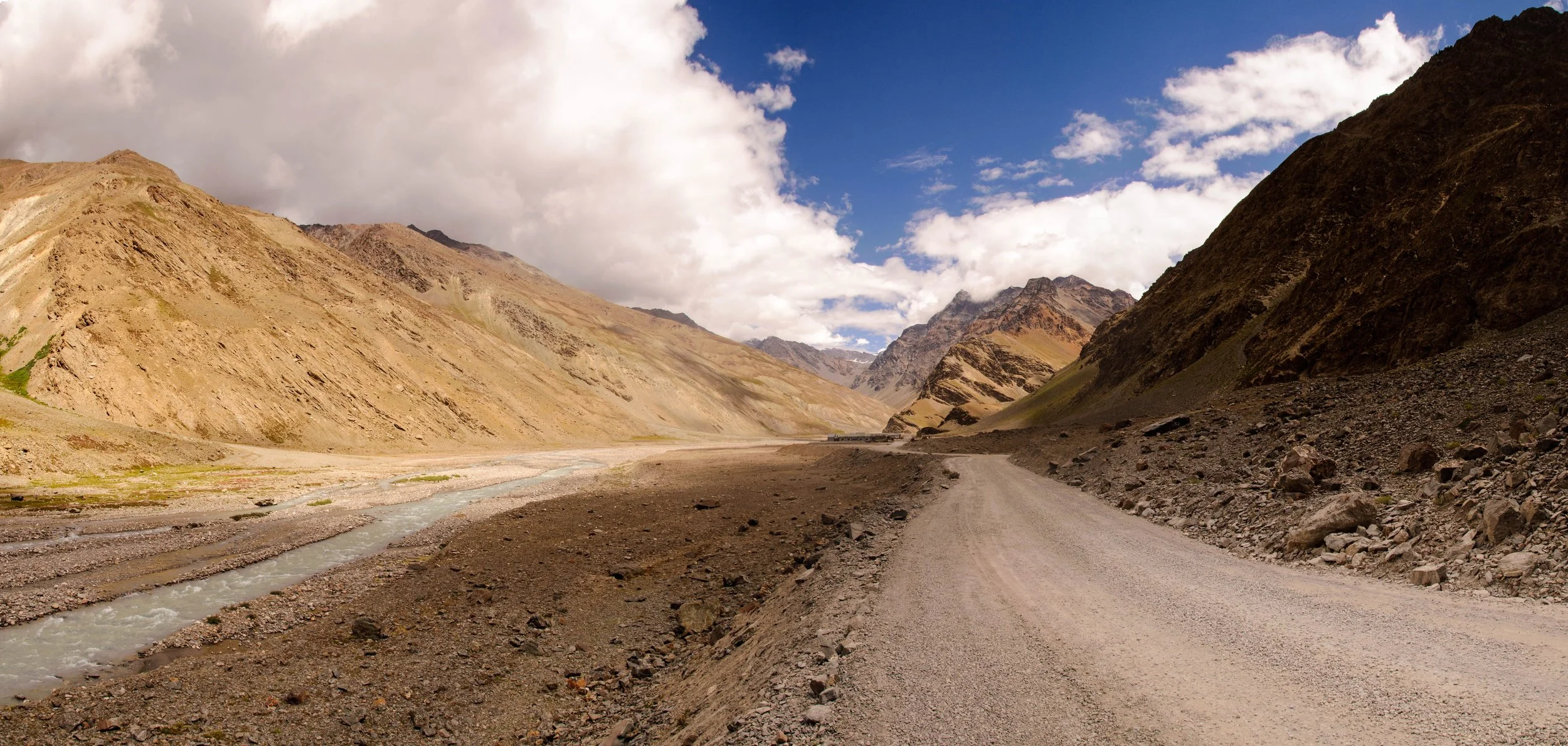 Dirt road winding through a mountainous desert landscape with rocky slopes on either side, under a partly cloudy sky.