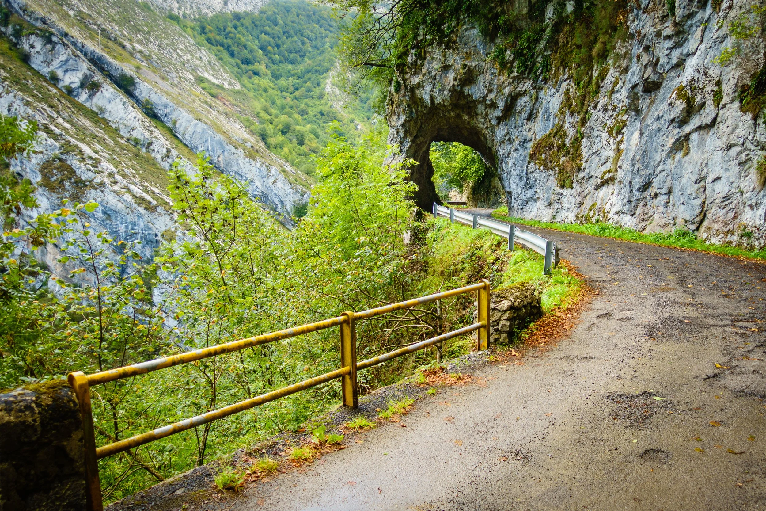 A narrow winding road through a rocky, forested mountain area with a natural rock arch formation overhead and metal guardrails on the edge of the road.