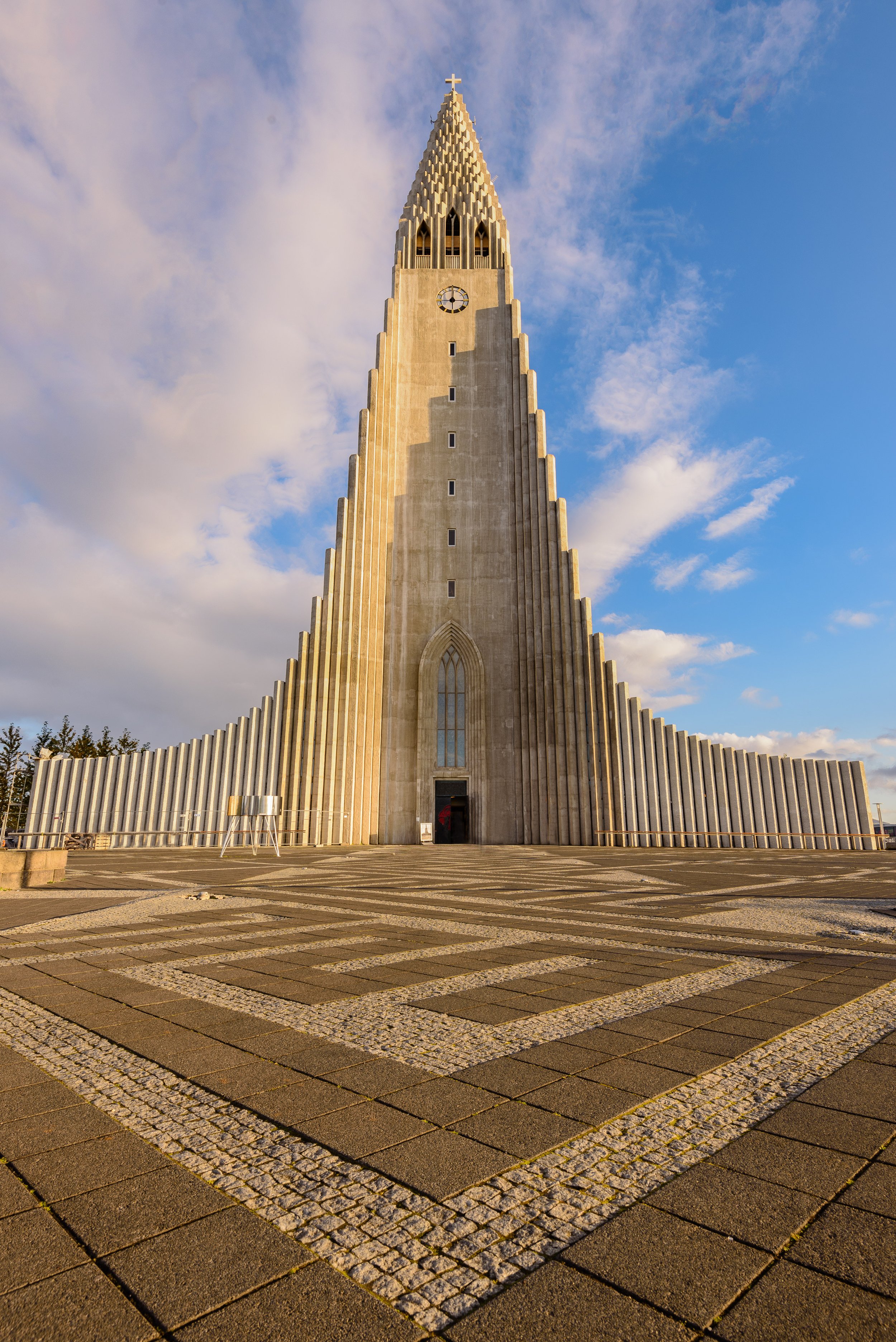 Hallgrímskirkja is a Protestant Lutheran (Church of Iceland) parish church in Reykjavík, Iceland, tall and narrow, with a clock near the top, set against a partly cloudy sky. The ground features a geometric pattern of paving stones.