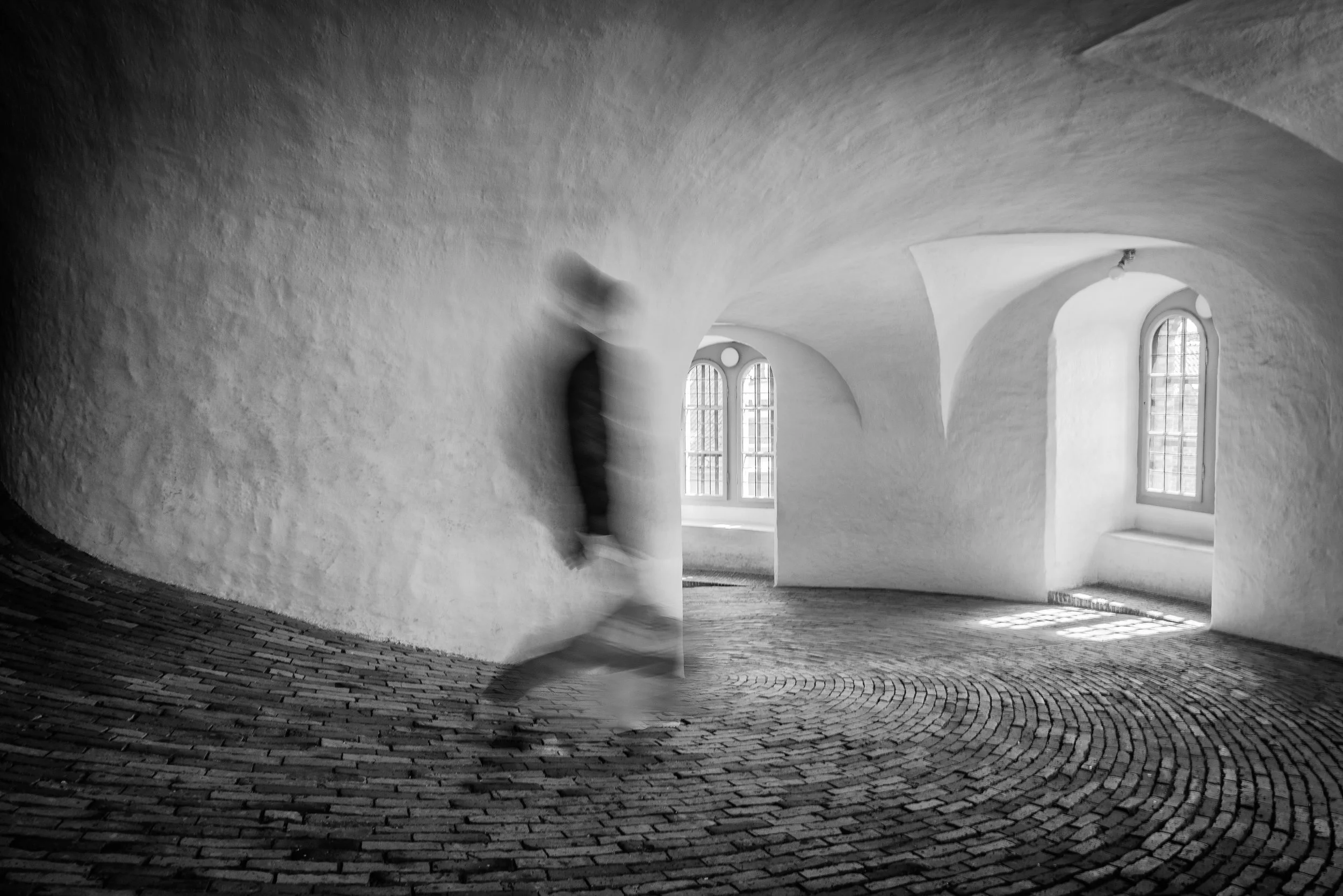 A black and white photo of a person walking through an art gallery with white, curved walls and cobblestone floor, with windows letting in natural light.