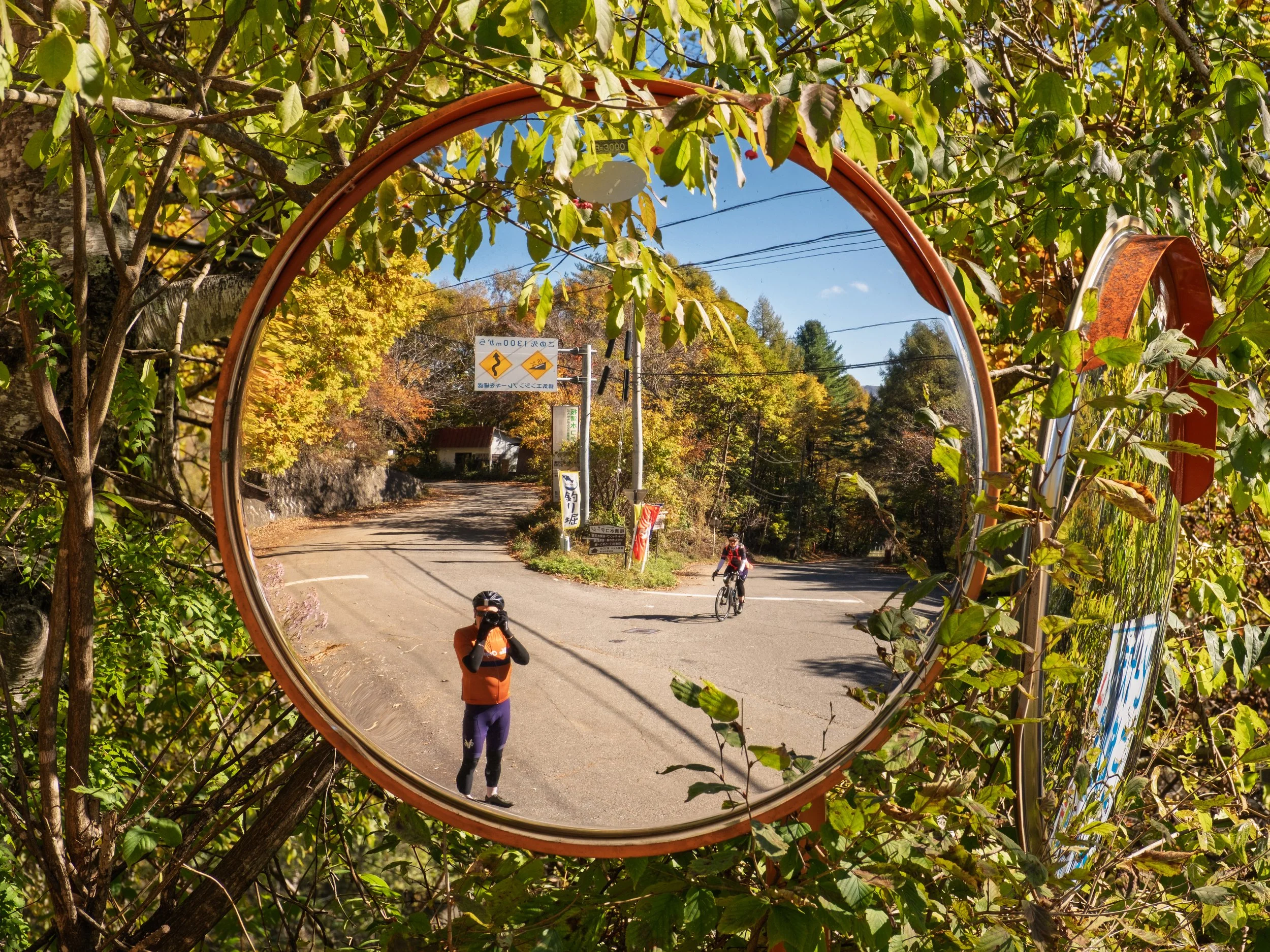 Reflection of a person taking a photo of two cyclists in a convex traffic mirror surrounded by green foliage on a sunny day.
