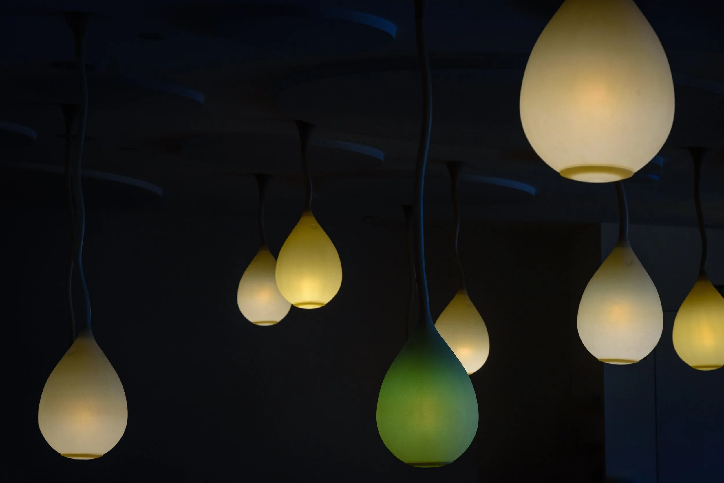 Multiple hanging teardrop-shaped pendant lights with a warm glow, suspended from a dark ceiling. One light has a green hue, while the others are yellowish.