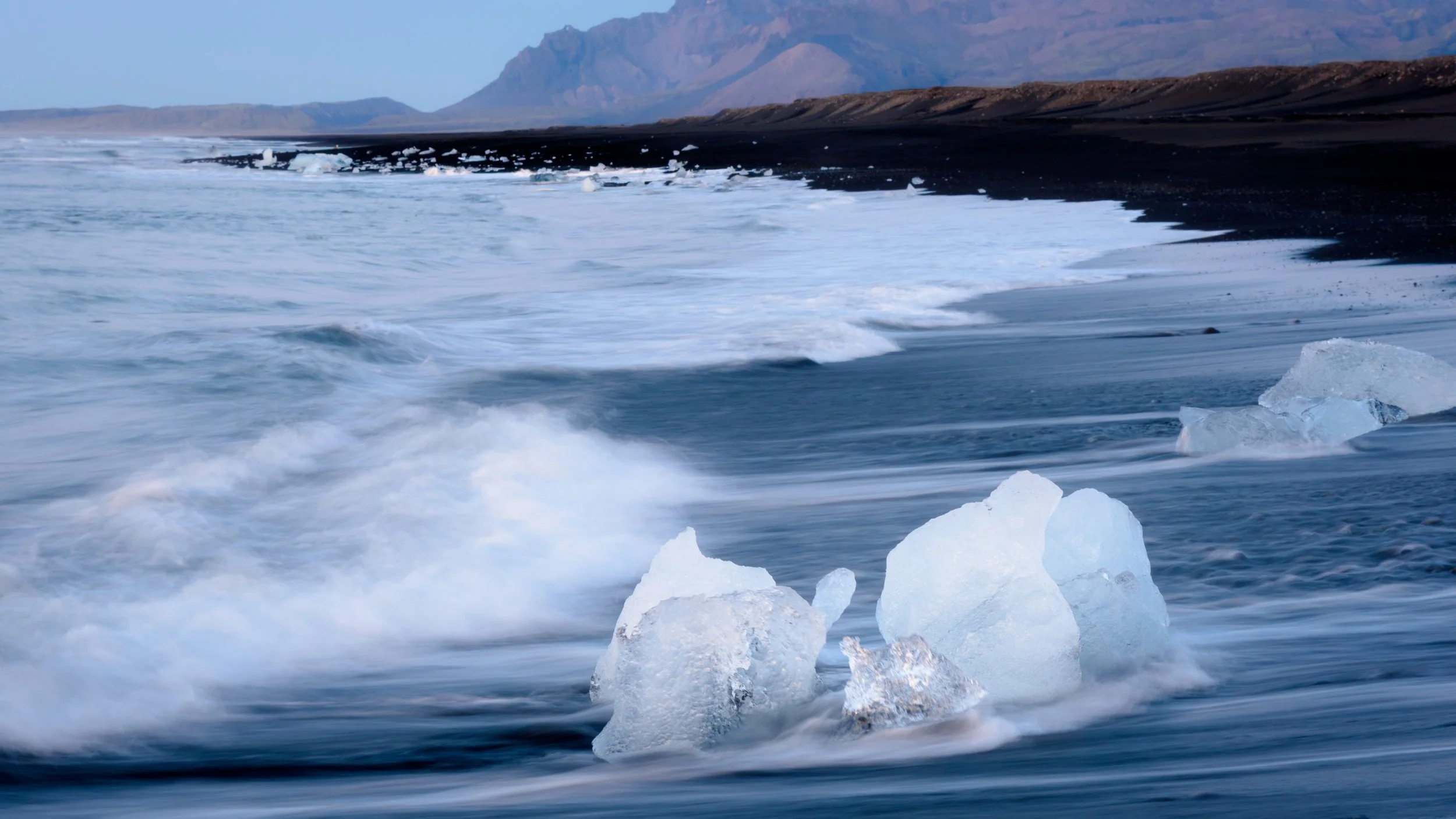 Ice chunks on a shoreline with ocean waves and mountainous landscape in the background.
