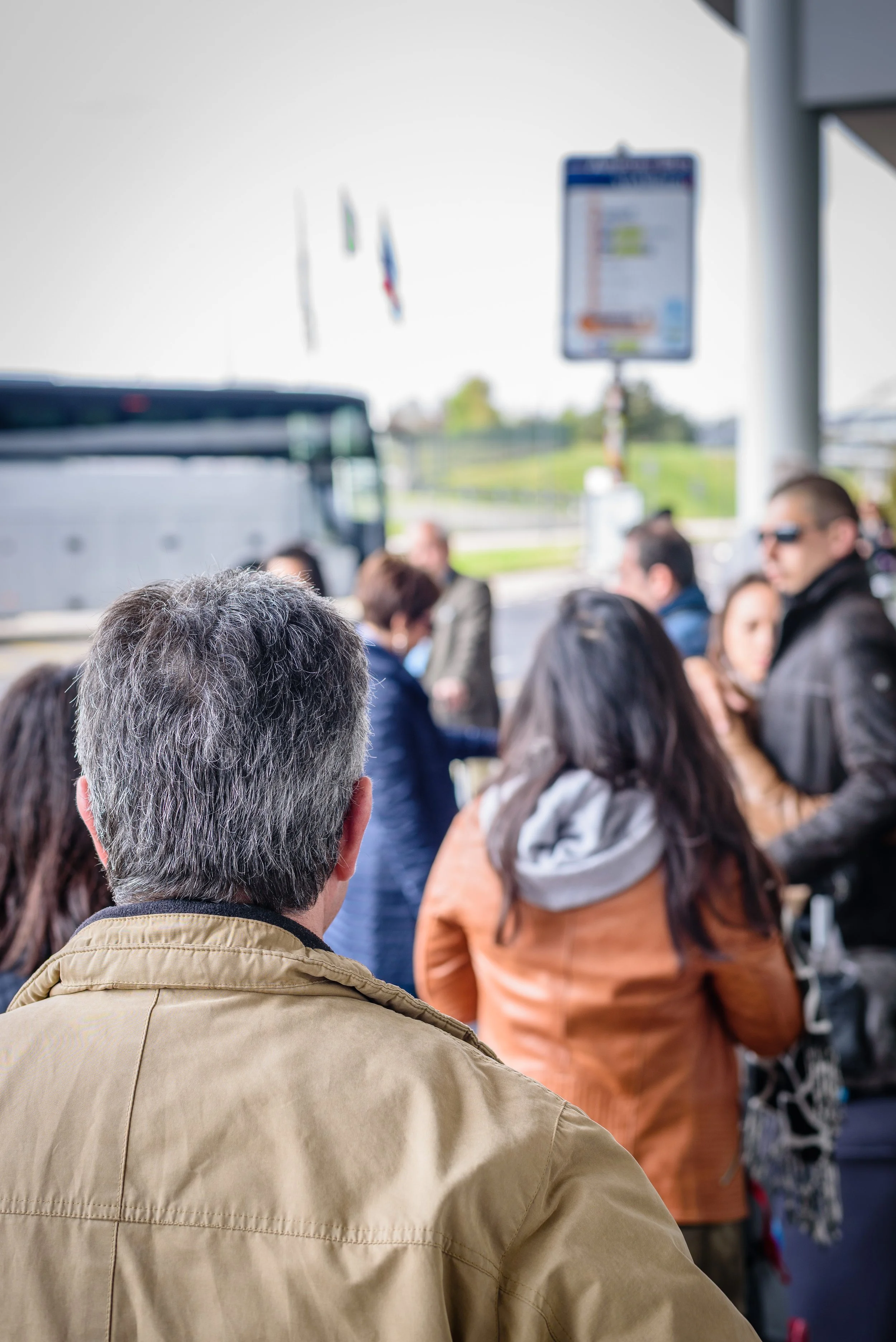 A man with gray hair wearing a tan jacket is waiting in line with a crowd of people outside, likely at a bus or transportation terminal. In the background, there's a bus and people standing near a sign near a building.