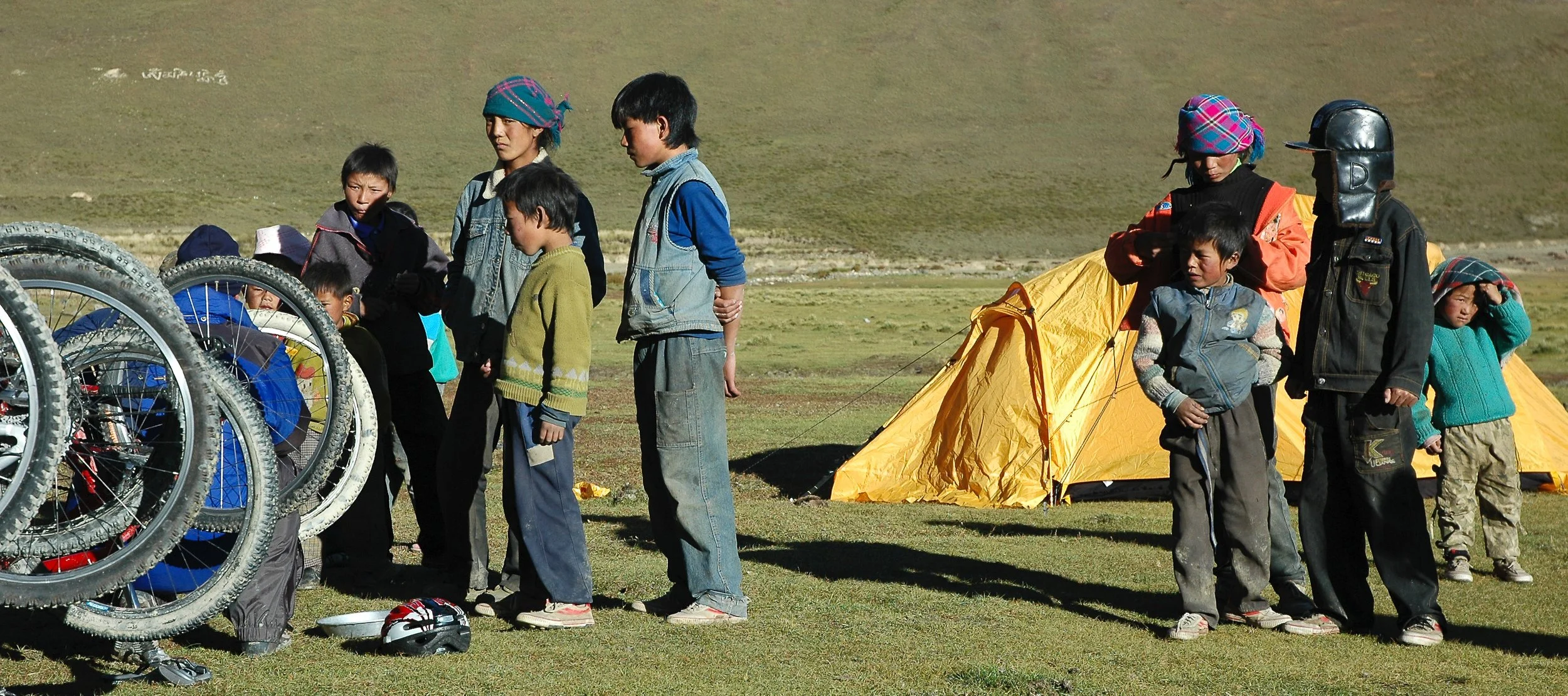 Group of children and adults standing outdoors near a yellow tent, with bicycle wheels in the foreground and a hill in the background.