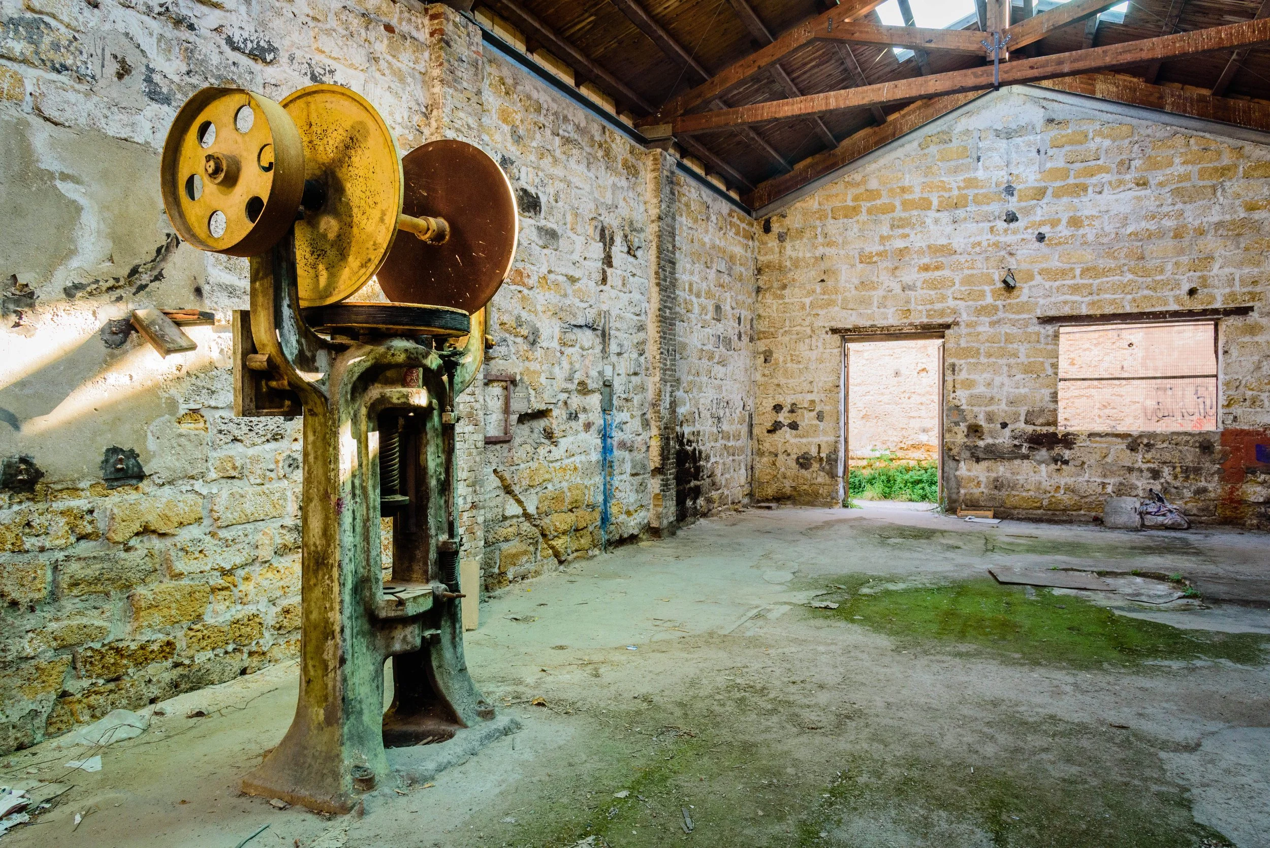 An old, rusted industrial meat slicer in a dilapidated warehouse with exposed brick walls, a dirt floor with patches of moss, and an open doorway showing an outdoor area.