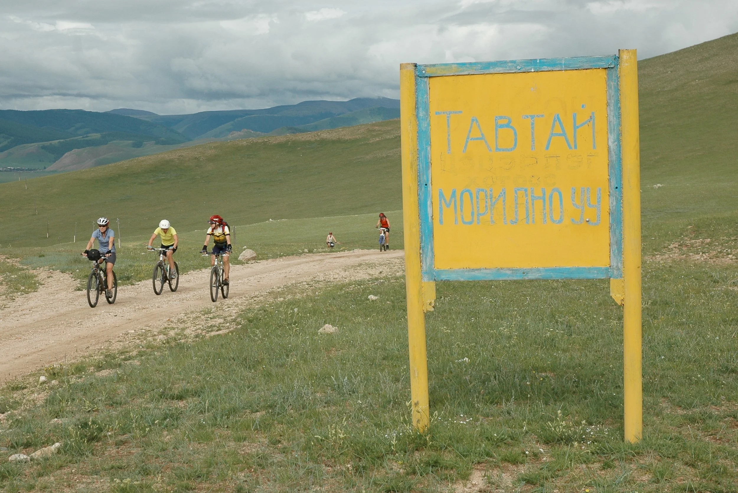 Bikers riding on a dirt trail across a green landscape with mountains in the background, and a yellow sign with blue text in Cyrillic script in the foreground.