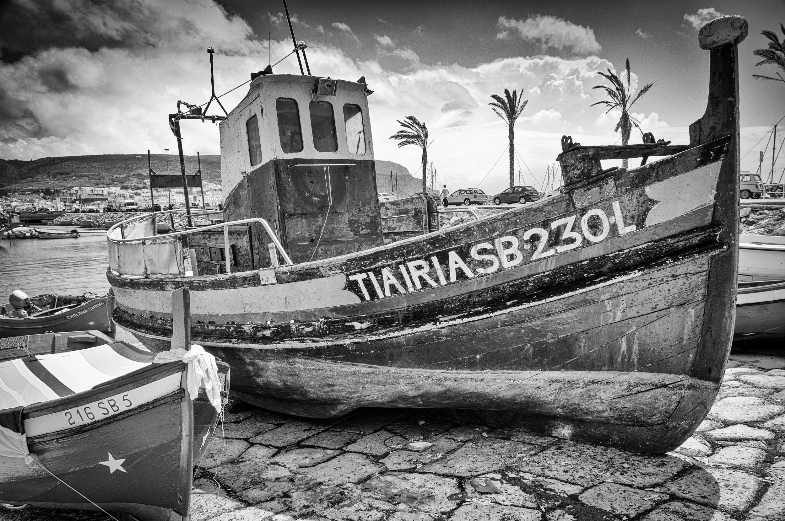 Old, weathered fishing boat with registration TIAIRIA SB 2301 on a harbor cobblestone surface, other smaller boats nearby, palm trees, cars, and mountains visible in the background under a cloudy sky.