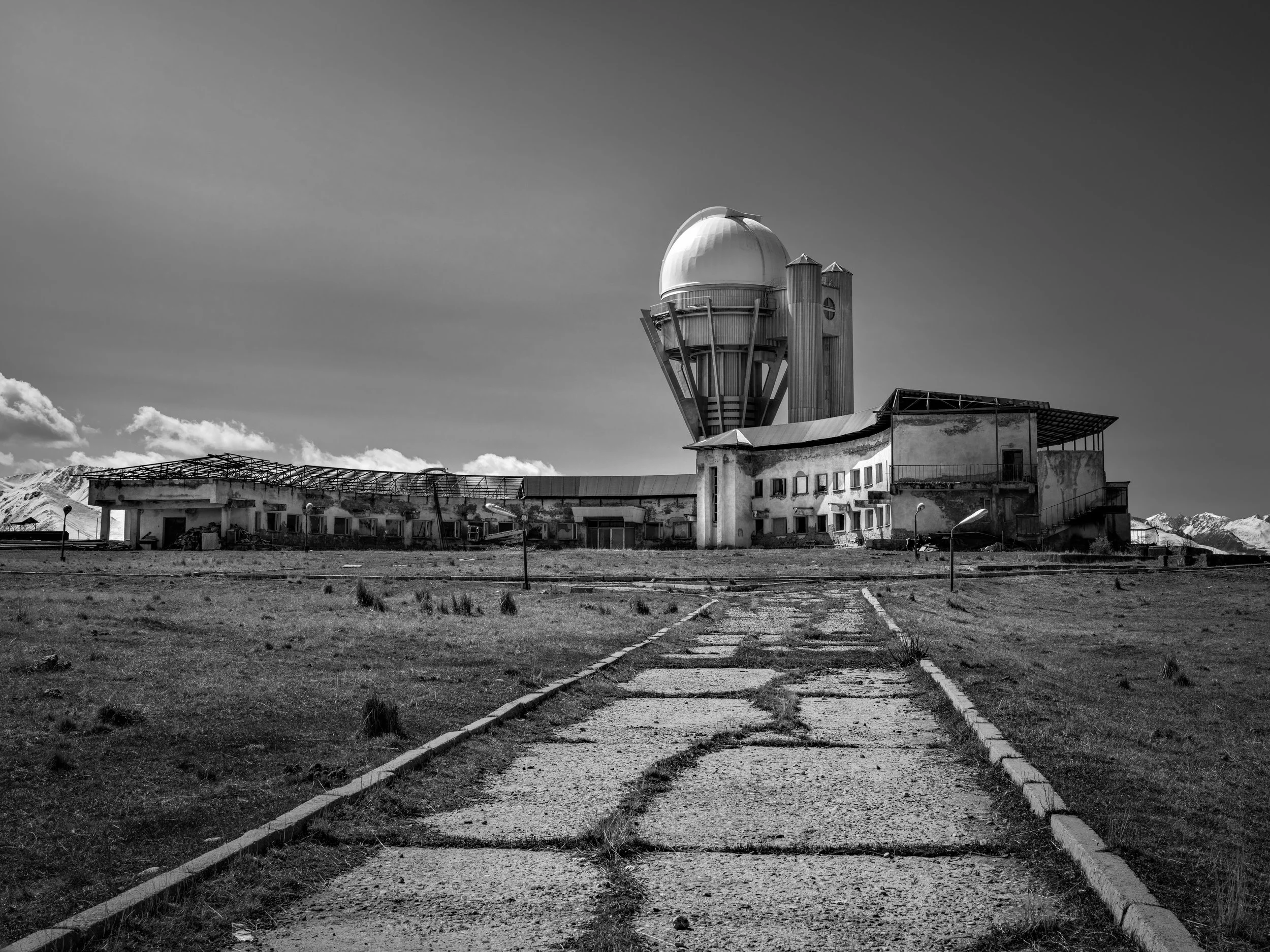 An abandoned, weathered observatory building with a large dome and surrounding structures, in a desolate landscape with snow-capped mountains in the background, under a cloudy sky.