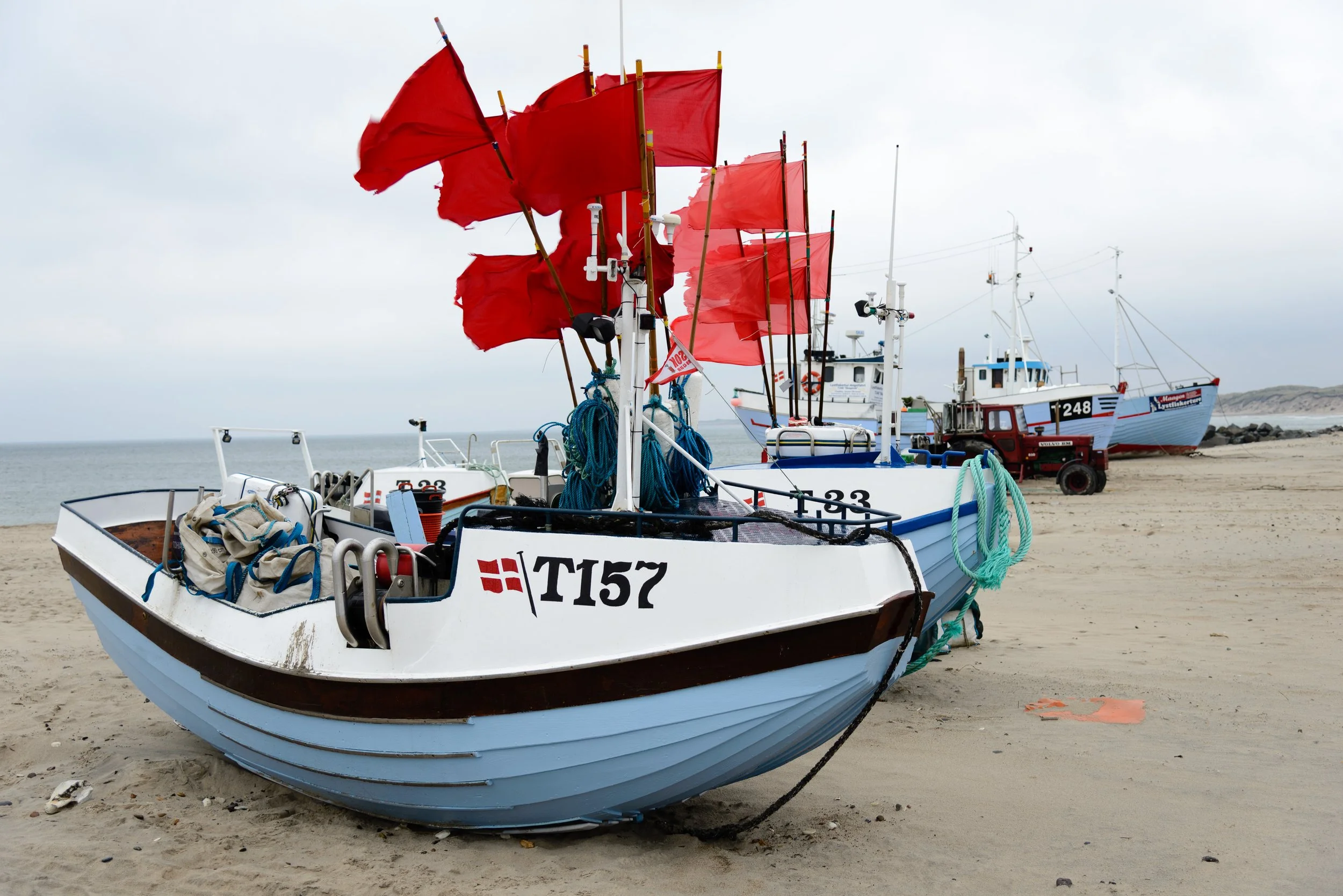 Several fishing boats with red flags resting on sandy beach, with the ocean in the background under cloudy sky.