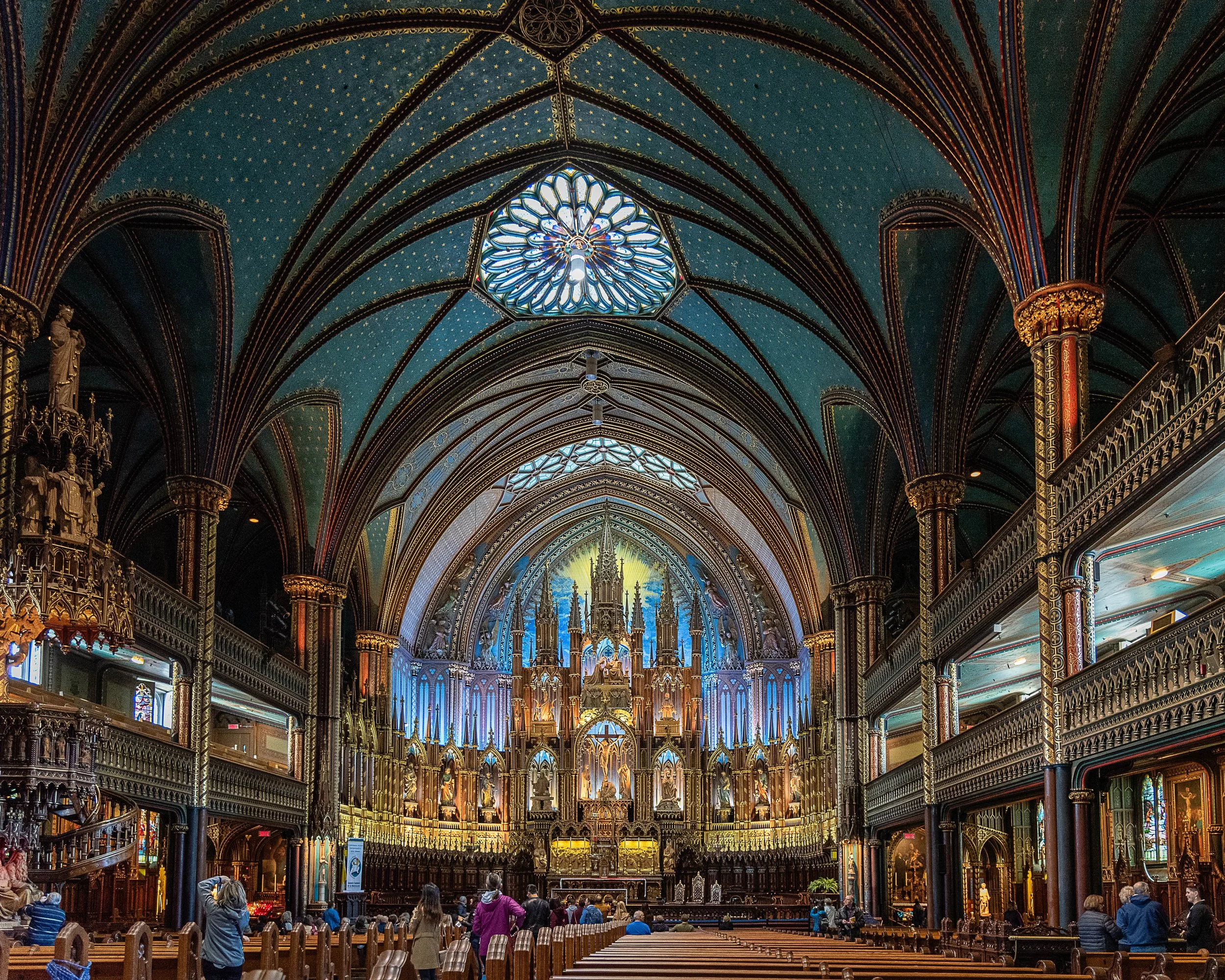 Interior of a grand church or cathedral with detailed stained glass windows, ornate gold and wood accents, and people seated and standing.
