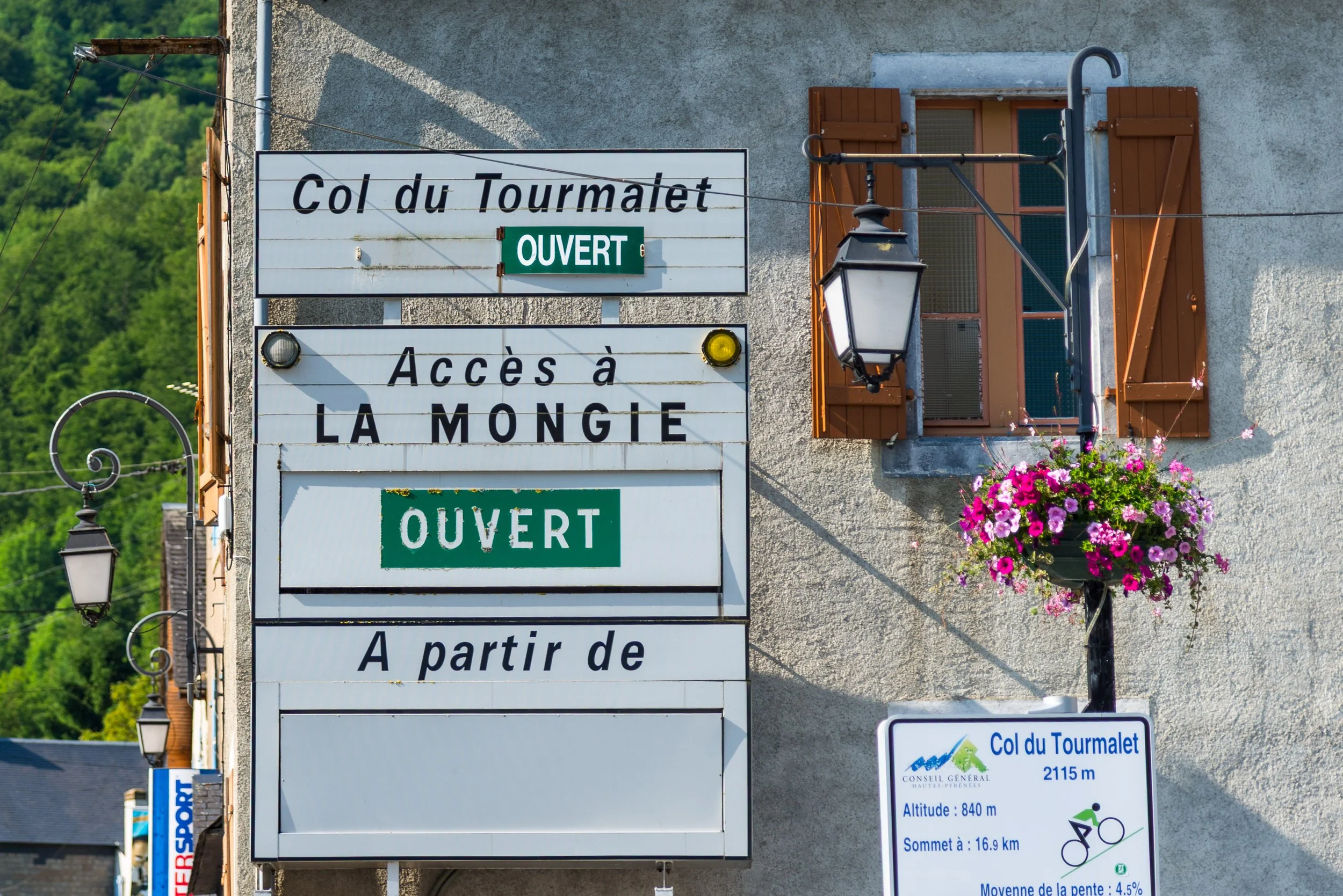 Signage for Col du Tourmalet, showing it is open, with access to La Mongie, a street lamp, a window with wooden shutters, and a flower basket with pink and purple flowers, set on a gray building exterior.