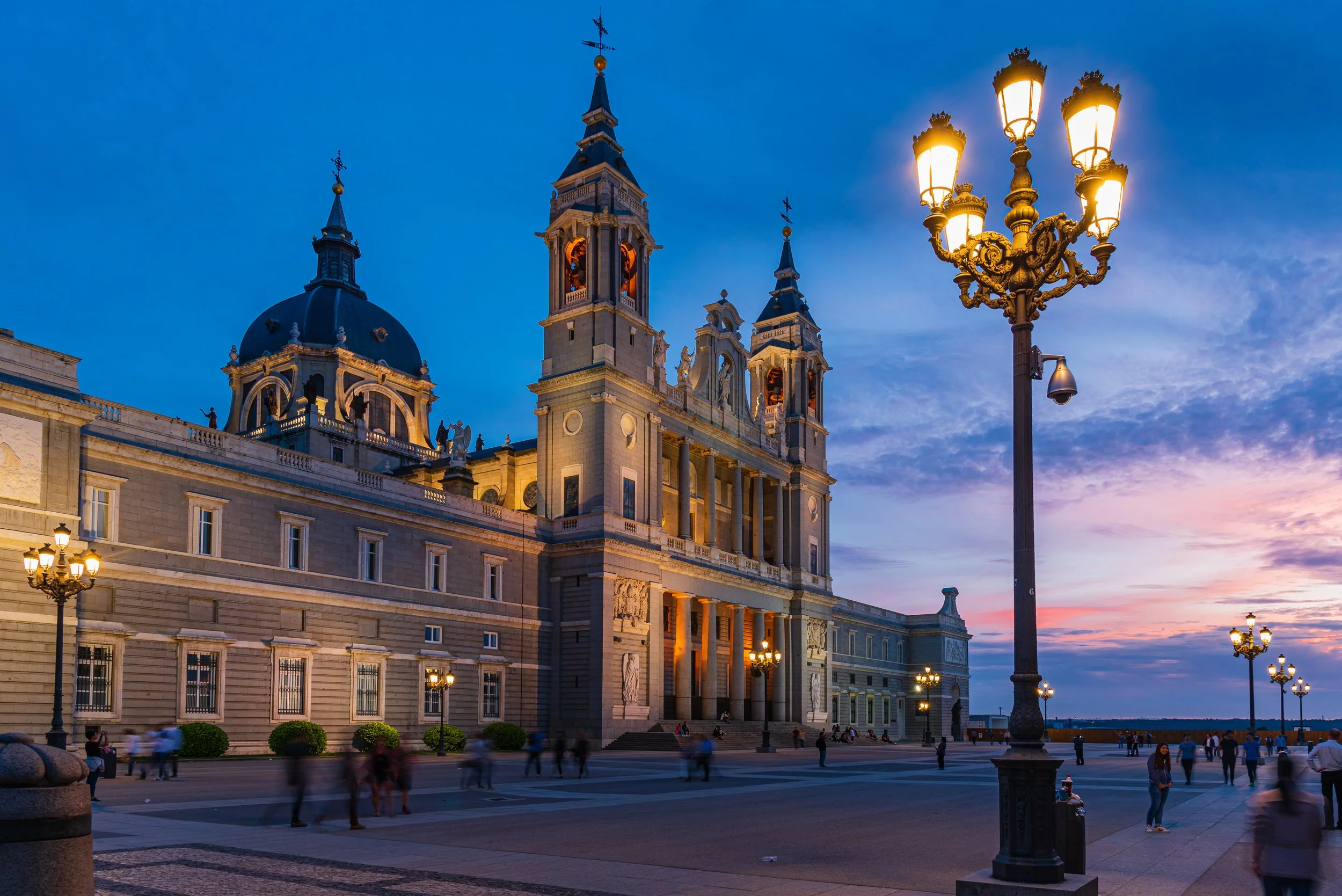 Exterior view of a grand historic cathedral with illuminated street lamps and people walking in the evening