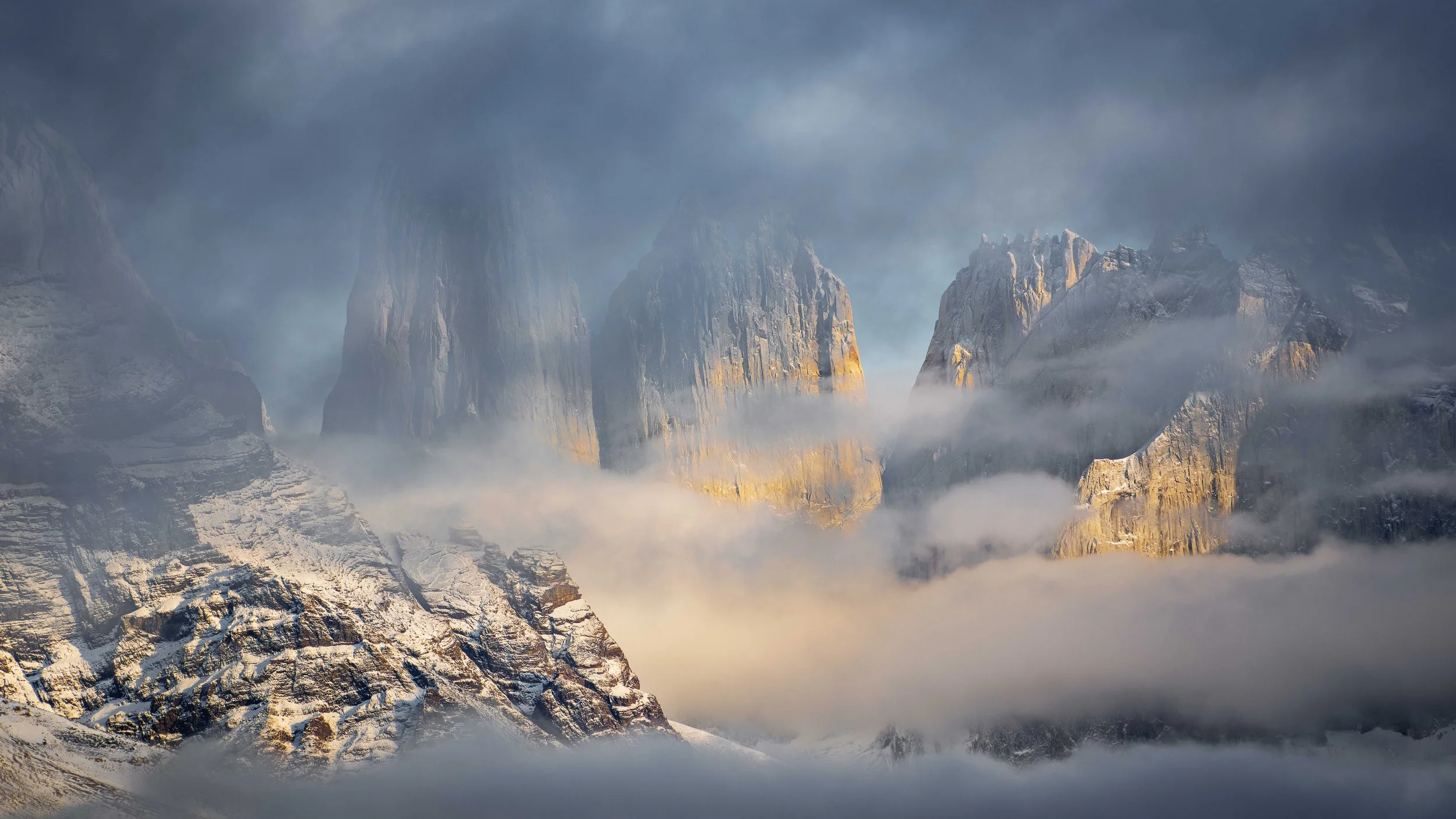 Snow-covered mountain peaks shrouded in clouds with a blue and gray sky in the background.