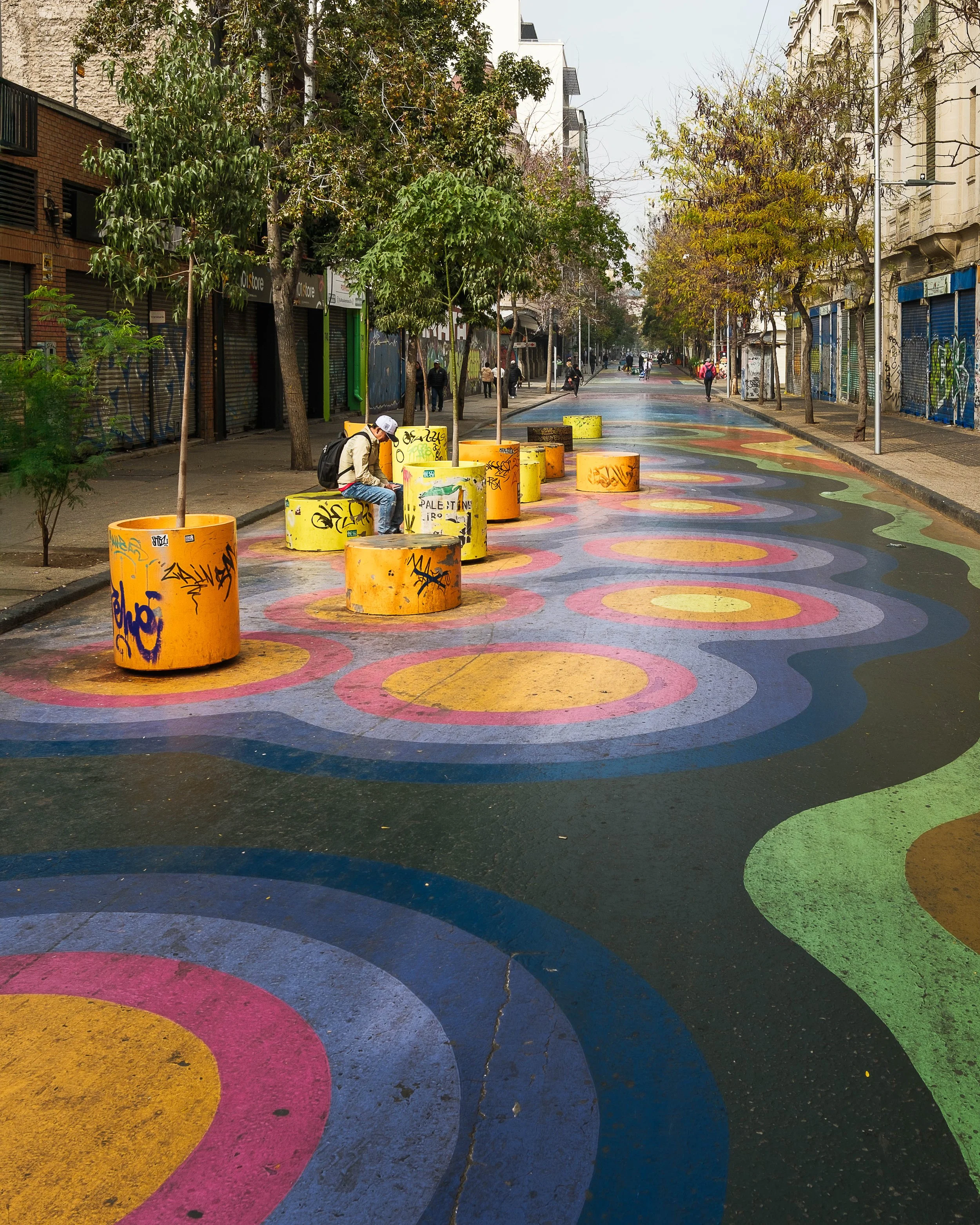 Colorful street with painted circular patterns, large yellow planters, trees, and a few people walking or sitting, in an urban area with buildings on both sides.