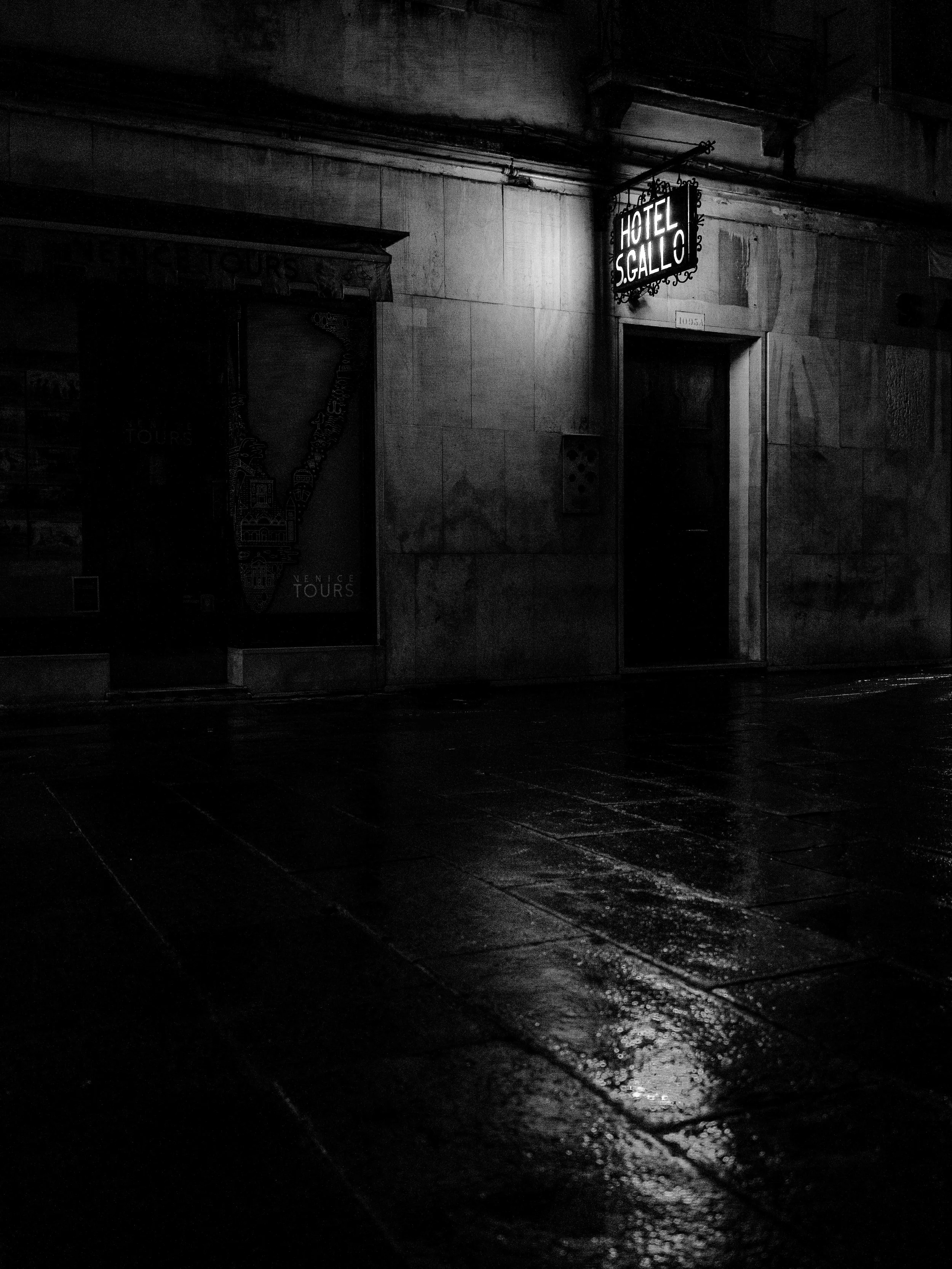 Nighttime street scene featuring a hotel sign reading 'Hotel S. Gallo' illuminated above a closed door, with wet pavement reflecting light, and a poster advertising Venice tours on the wall.