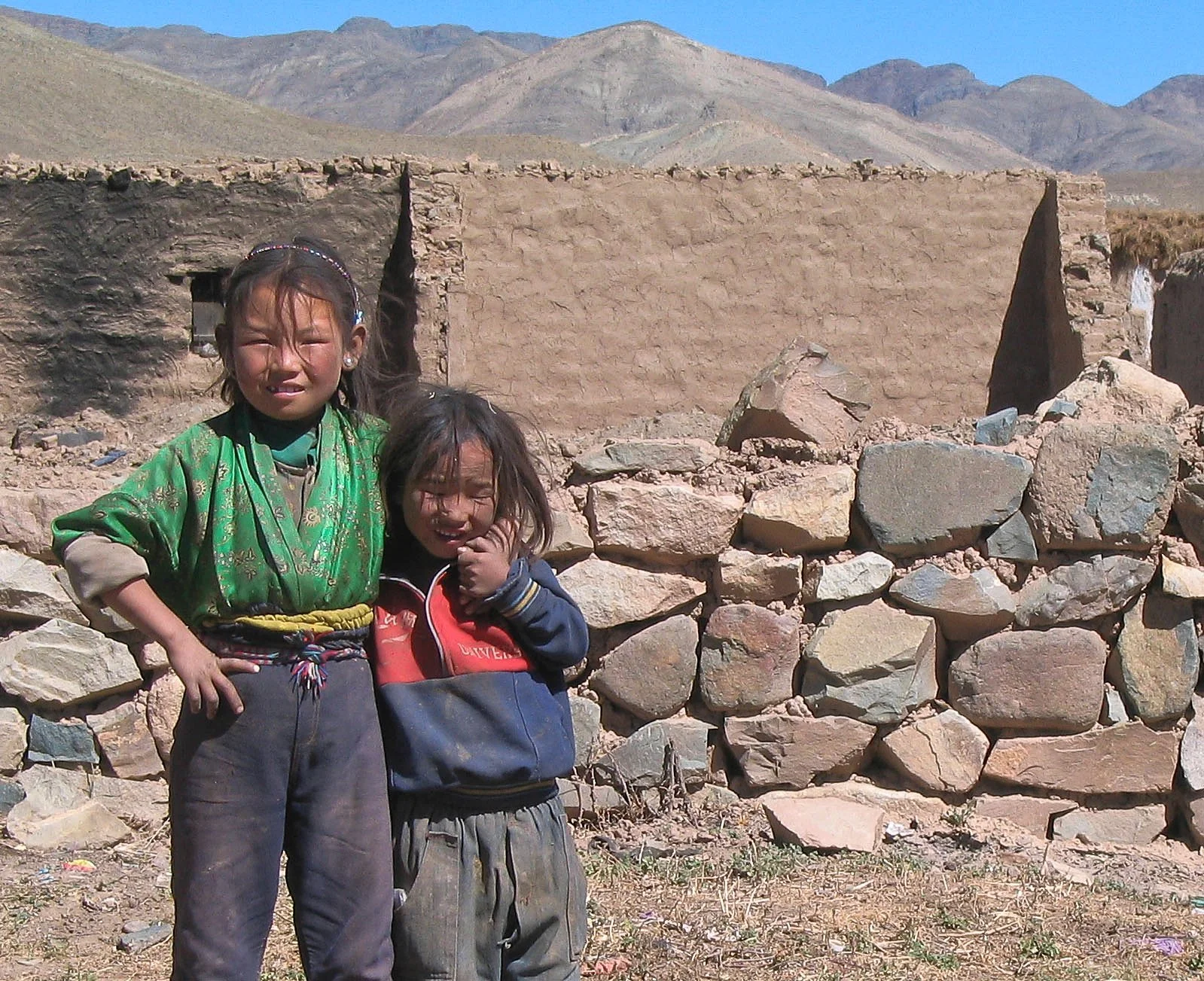 Two young children, a girl in a green jacket and a boy in a blue and red jacket, standing outside in a mountainous desert area with a partially built stone wall and an adobe structure in the background.