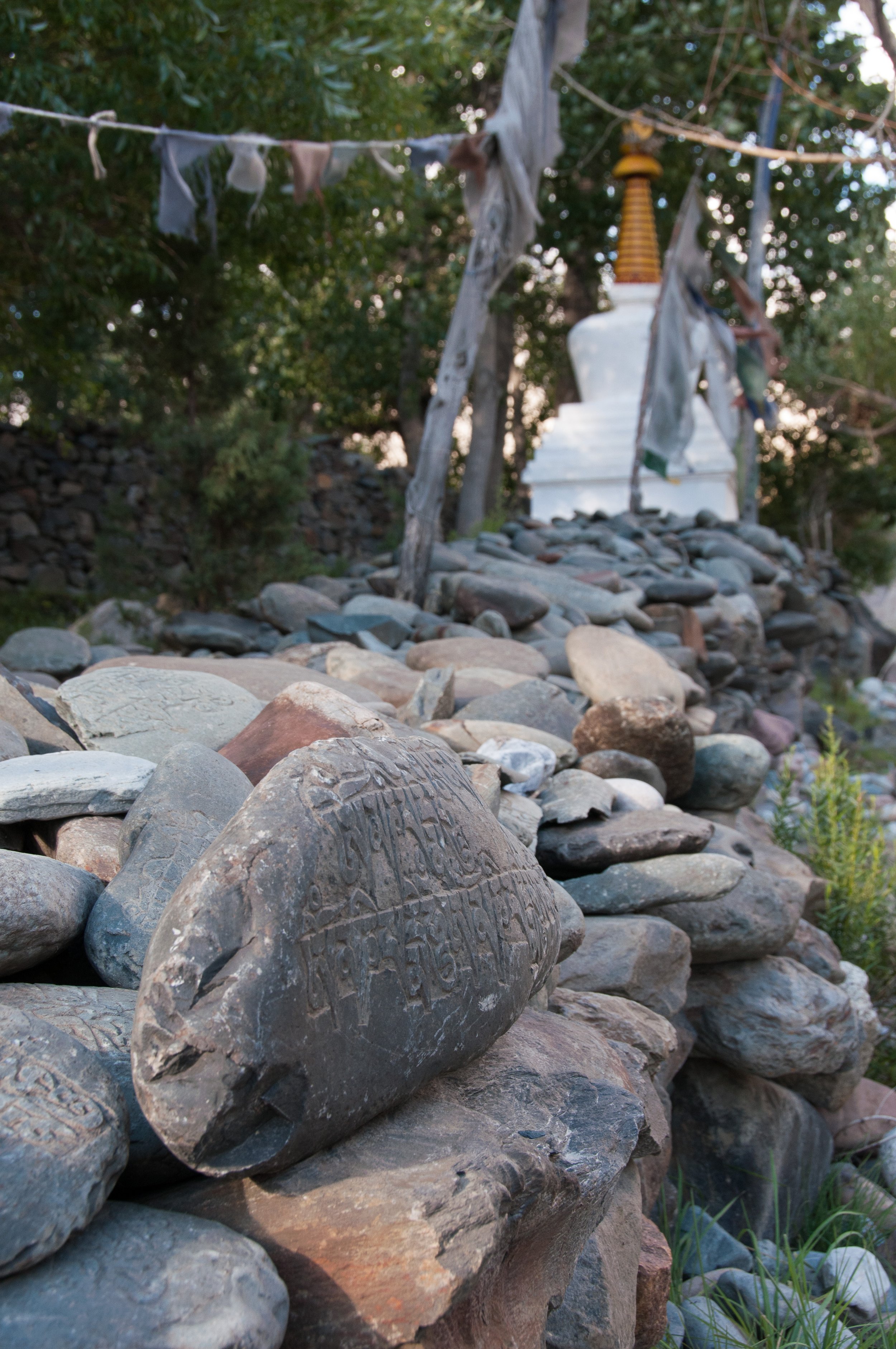 Close-up of carved stones on a rocky path leading to a white Buddhist stupa with prayer flags hanging nearby, surrounded by trees.