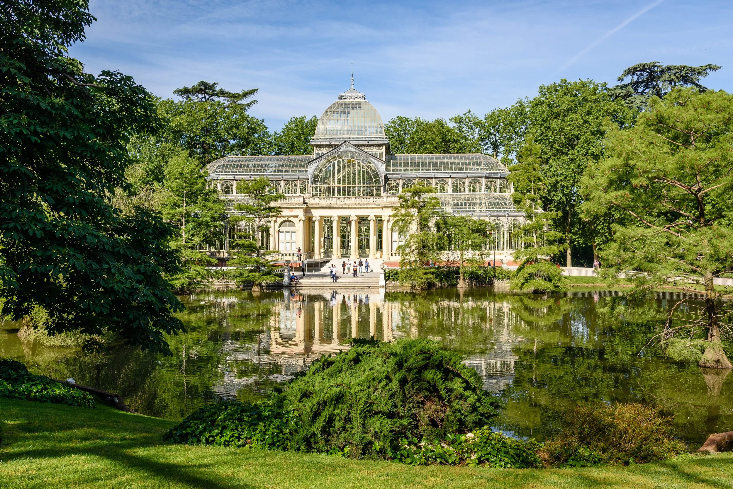A large glass greenhouse with classical architecture, featuring a prominent dome, surrounded by trees and a pond reflecting the building, with visitors standing on the steps in front.