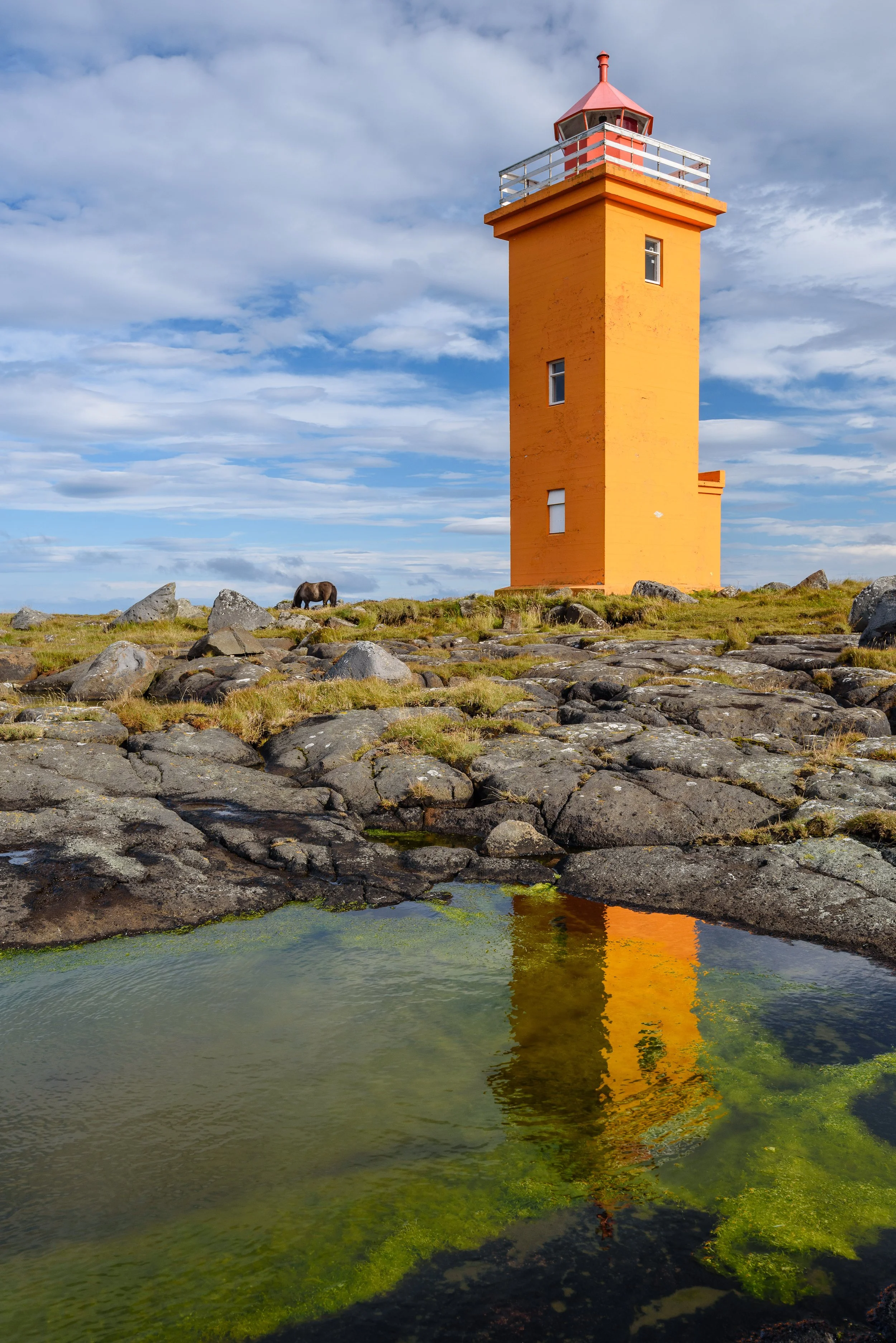An orange lighthouse on a rocky landscape near a small pond reflecting the lighthouse, with a blue sky and scattered clouds in the background.