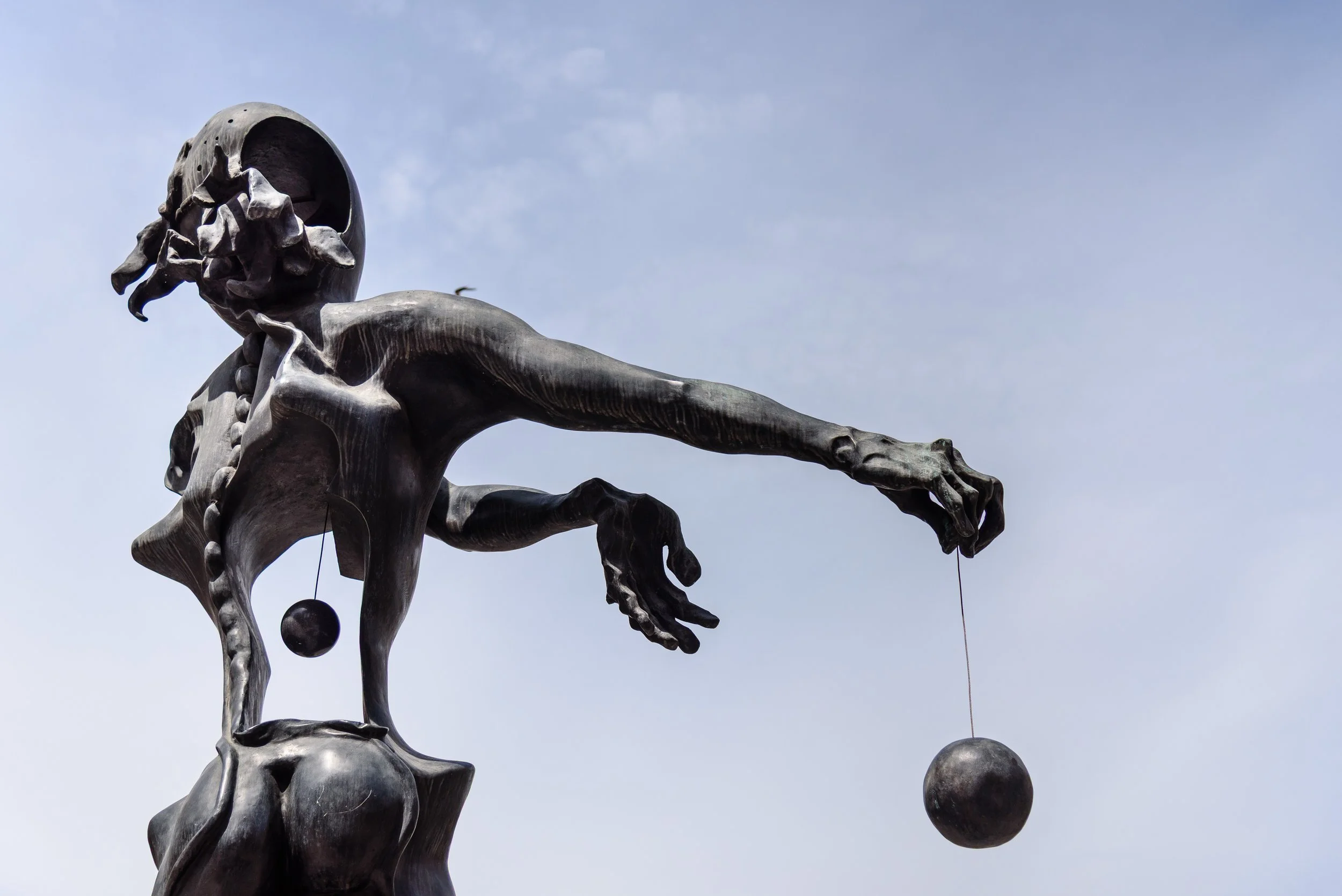 A metallic statue of a girl with a bob haircut and braided hair, holding a string attached to a ball, against a partly cloudy sky.