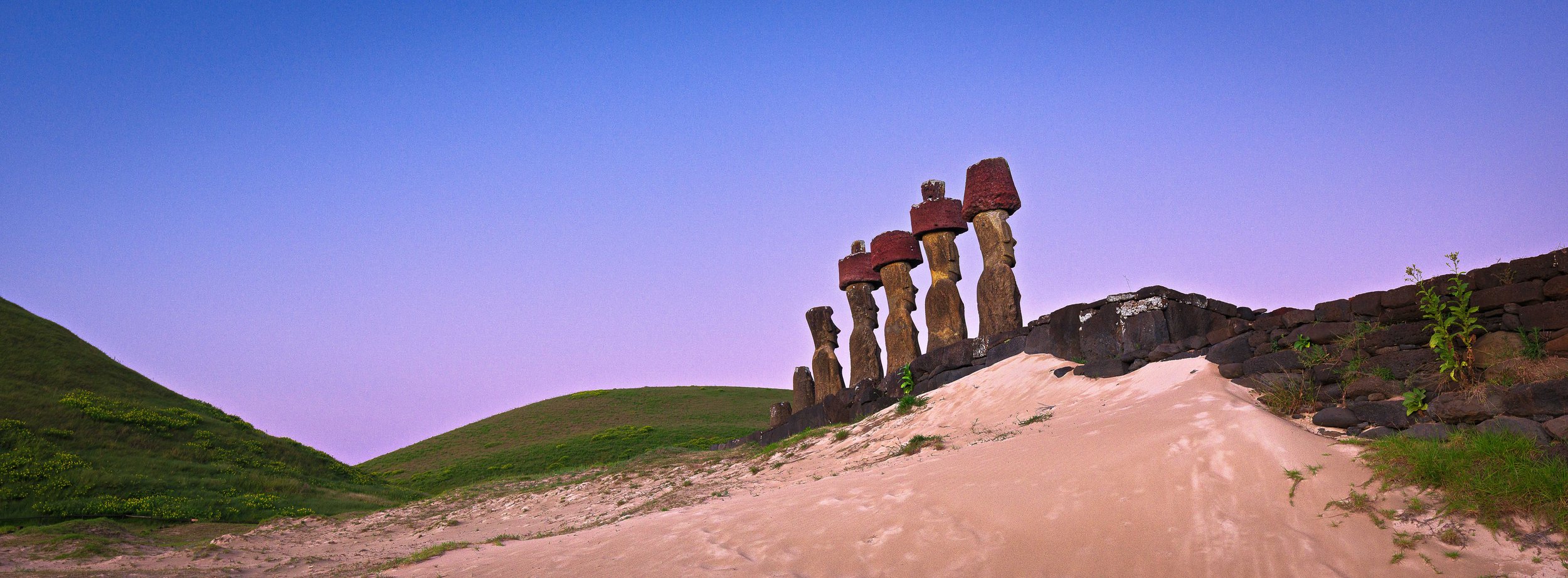Six Moai statues on a sandy beach with green hills under a pastel purple sky.
