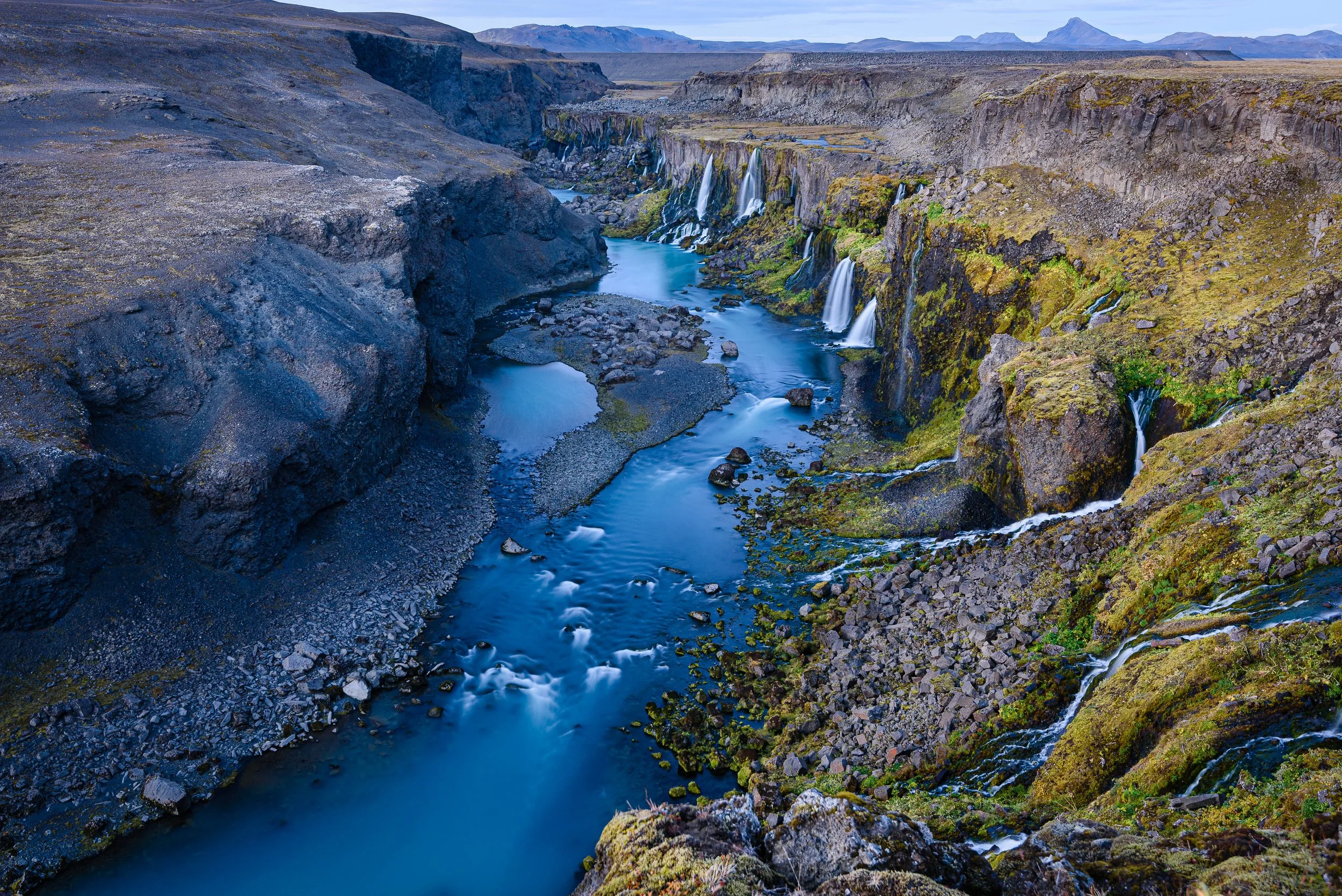 A scenic view of rugged cliffs and a river with multiple waterfalls in a volcanic landscape, likely in Iceland.