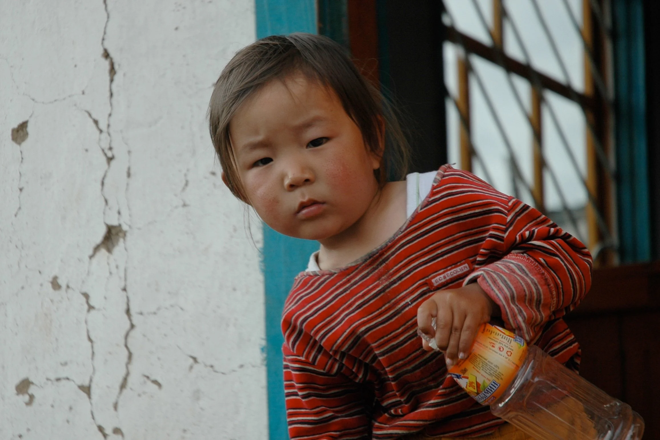 A young child with a serious expression, holding an orange plastic bottle, standing by a weathered white wall with cracks, and a window with blue bars in the background.