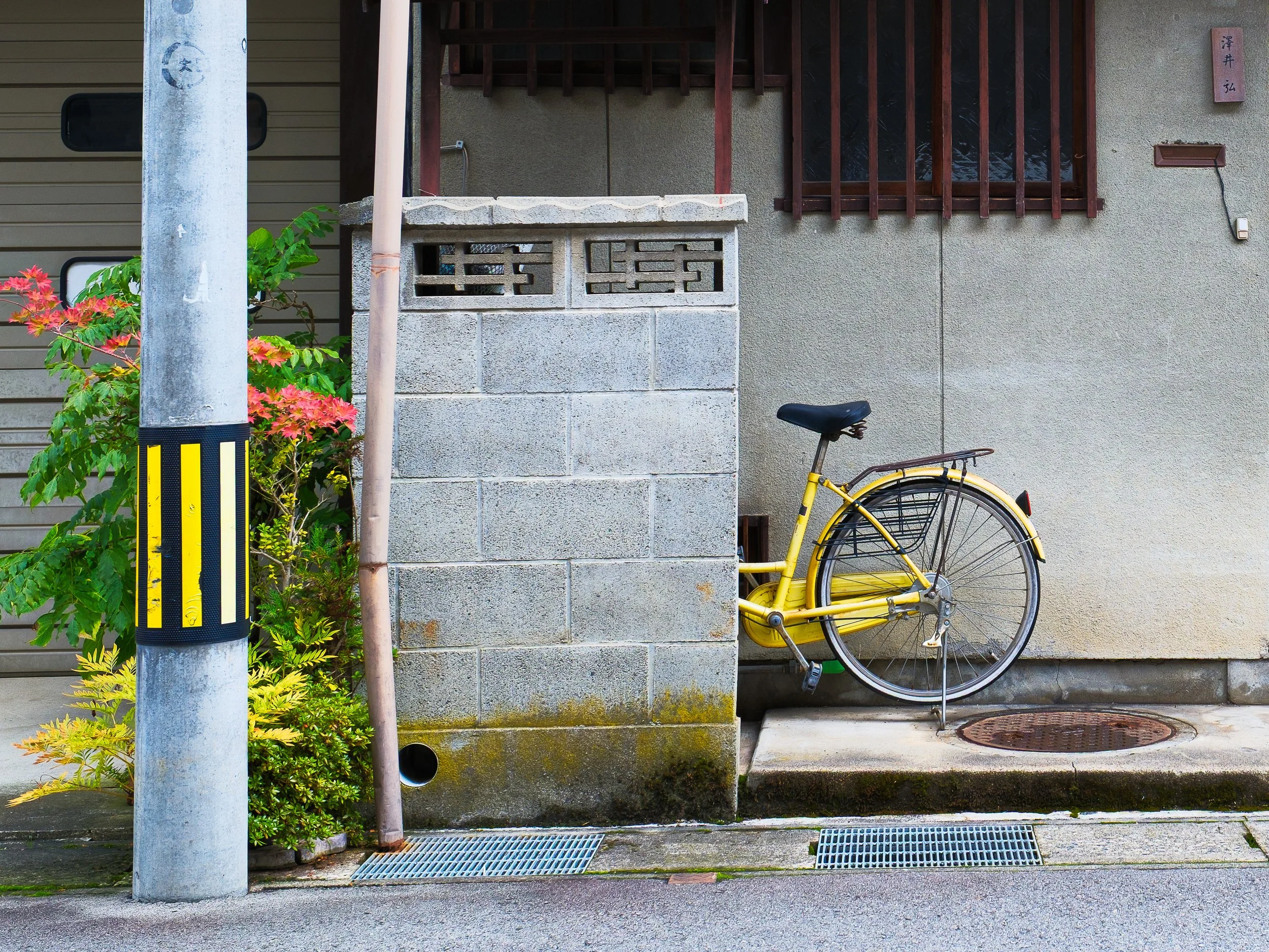 Yellow bicycle parked next to a concrete wall on a sidewalk, with a manhole cover and a drain, and a pole with yellow and black hazard markings in a residential alley.
