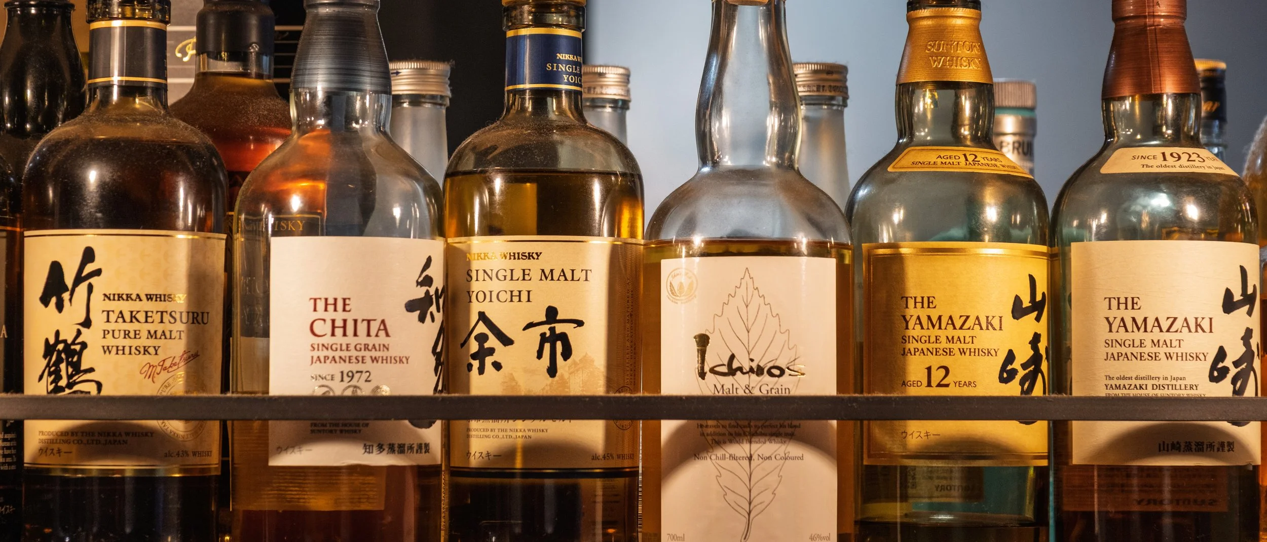 A shelf with various bottles of Japanese whisky and whiskey, including brands like Taketsuru, The Chita, Yoichi, Yamazaki, and Yaizu, labeled with different ages and types.