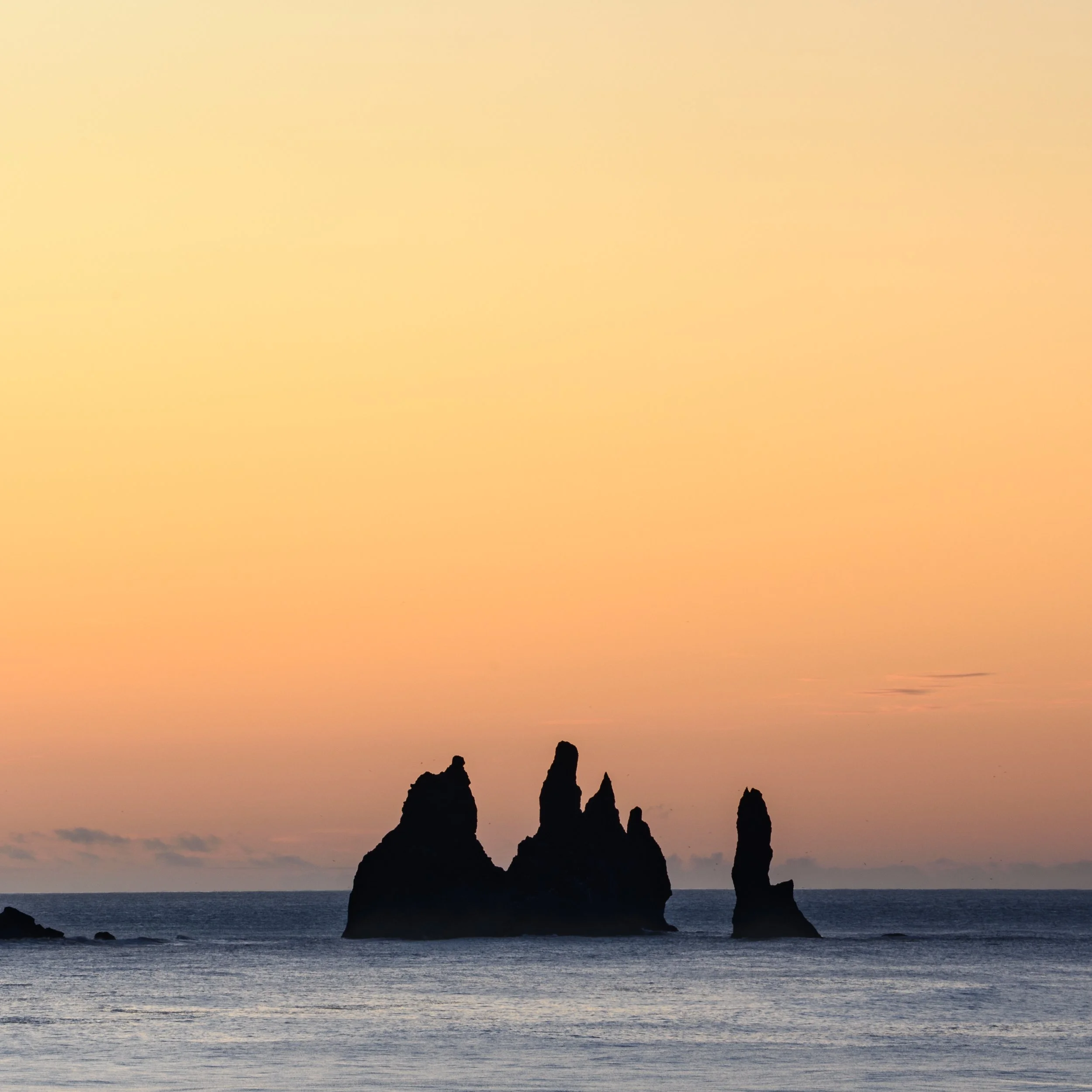 Silhouettes of tall rocks emerging from the ocean at sunset with a pastel orange and yellow sky.
