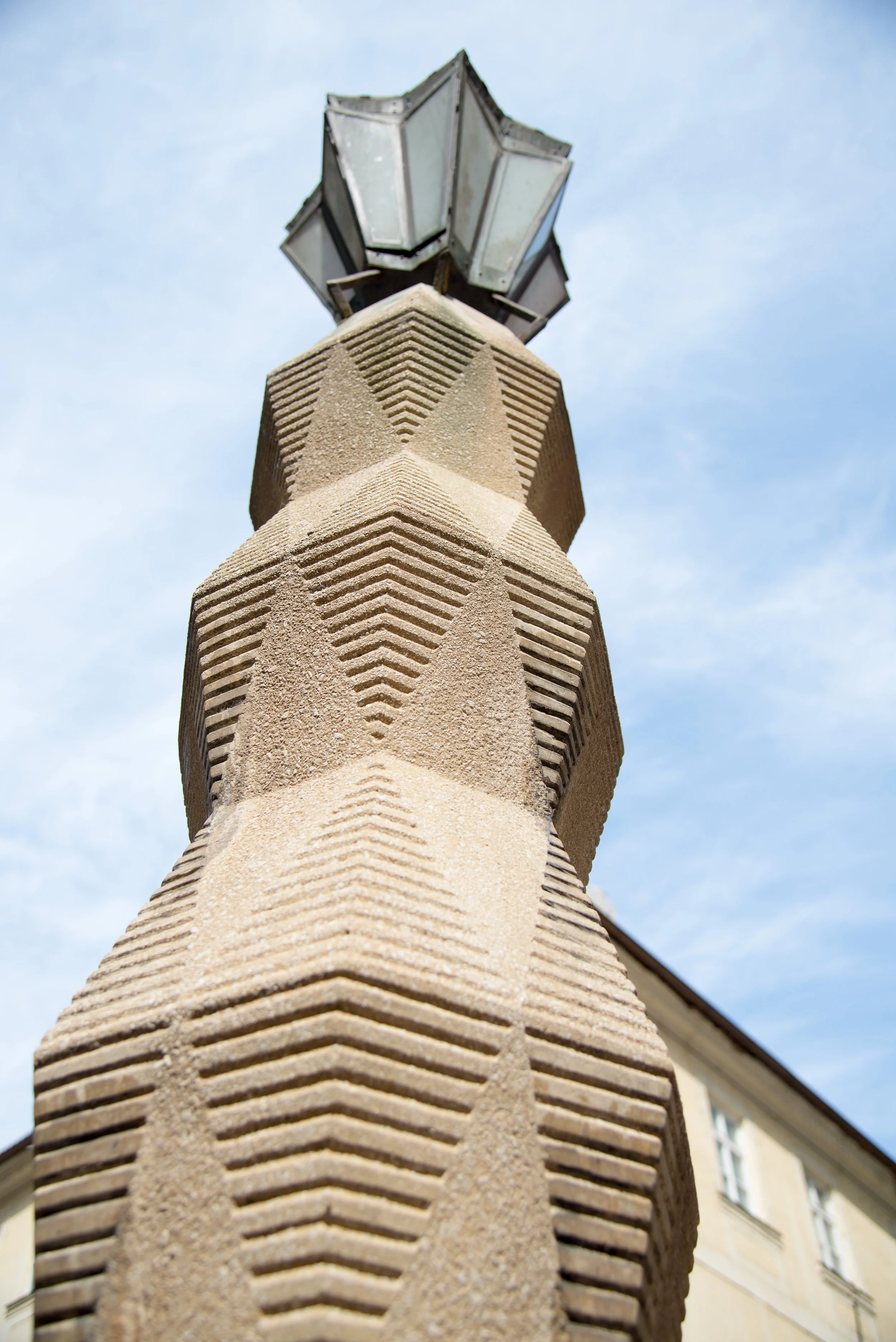Close-up of a decorative stone column with geometric patterns, topped with a vintage street lamp against a blue sky with a few clouds.