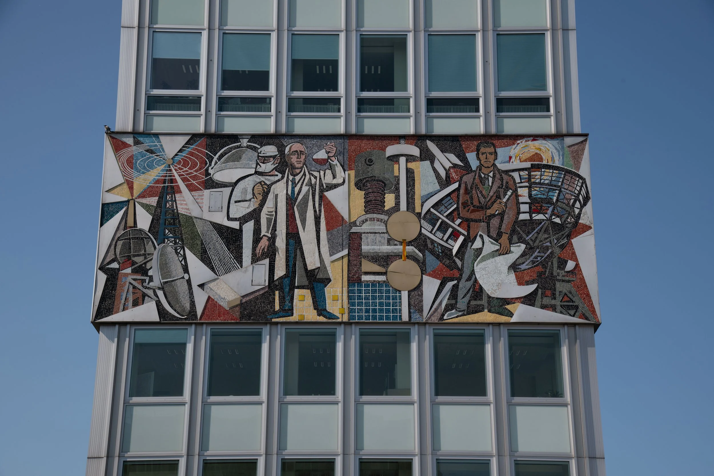 A large mural on the side of a building depicting scientists and technological themes, including a scientist with a test tube, a modern industrial scene, and scientific equipment, set against a vibrant geometric background.